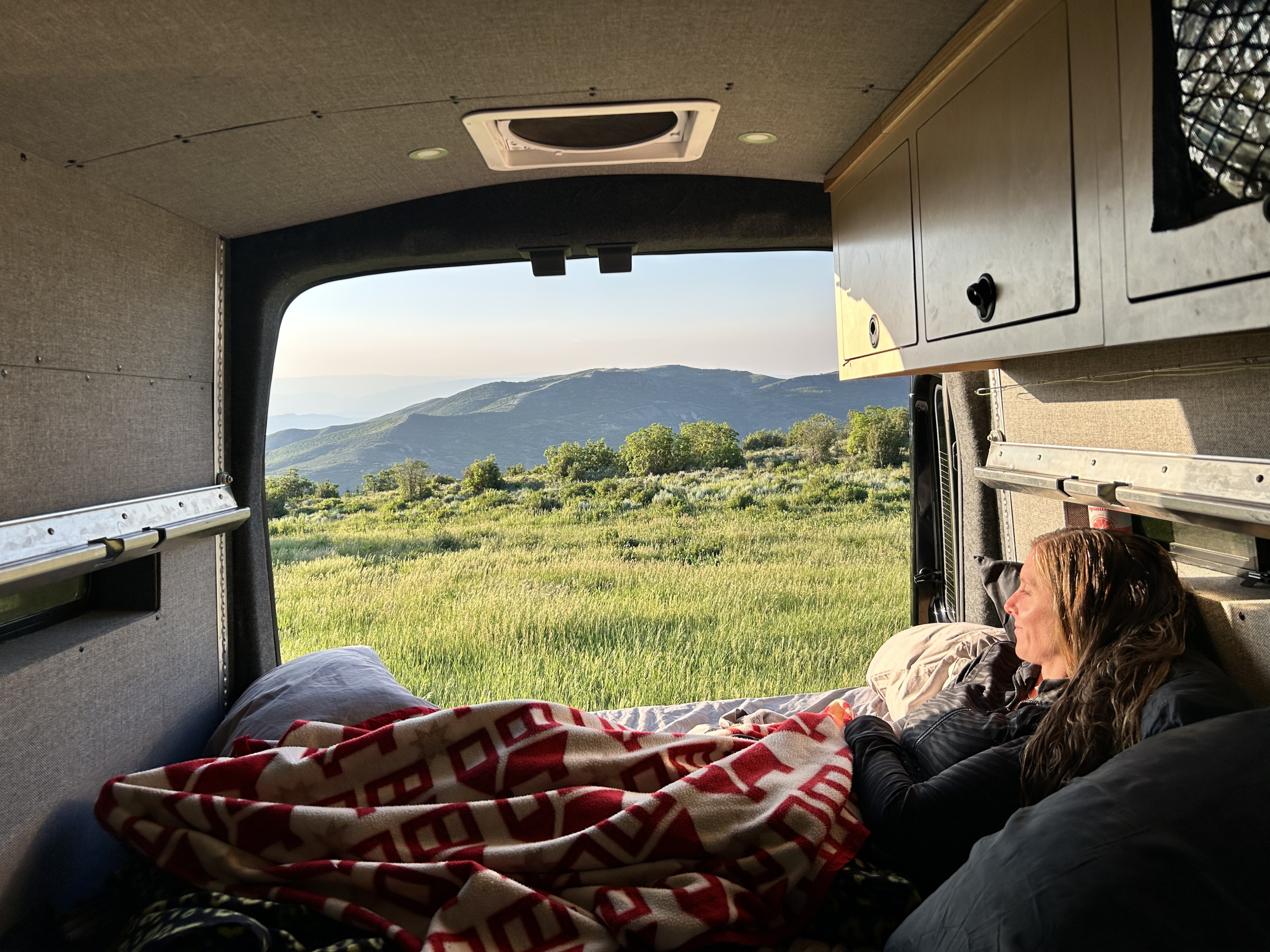 Relaxing in an rv rental camper van, a woman enjoys the mountain view. She's covered by a blanket, gazing at a grassy landscape.. Ford Transit 2023