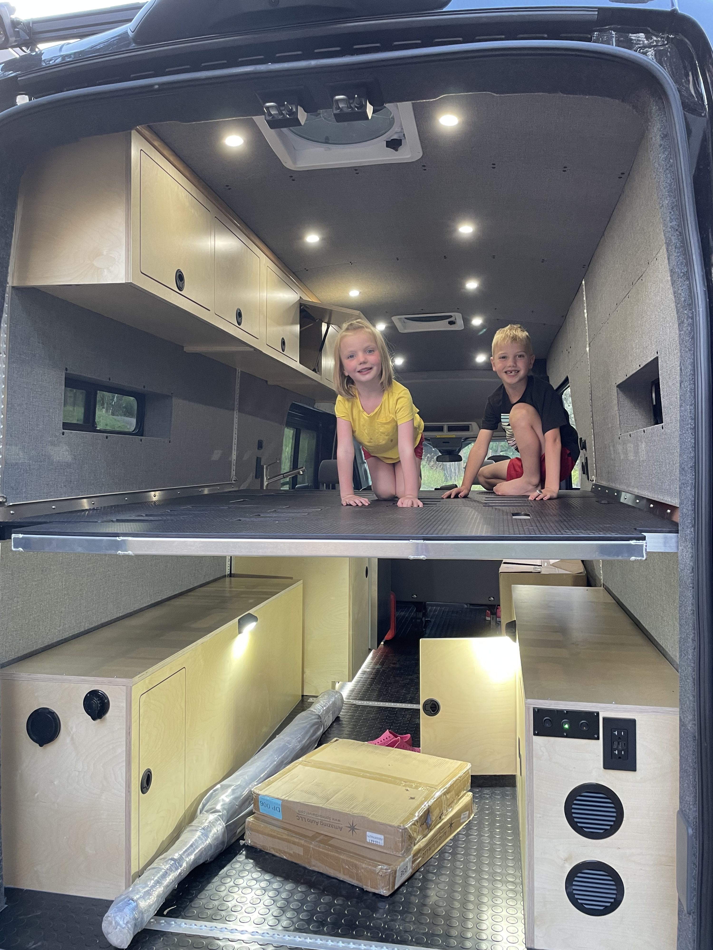 Two kids happily play on the elevated bed platform inside a custom RV rental. Modern van interior with storage, lights, and amenities.. Ford Transit 2023