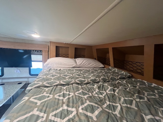 RV rental interior featuring a comfortable bed, overhead storage cubbies with nets, a TV, and a table.. Thor ACE 2015