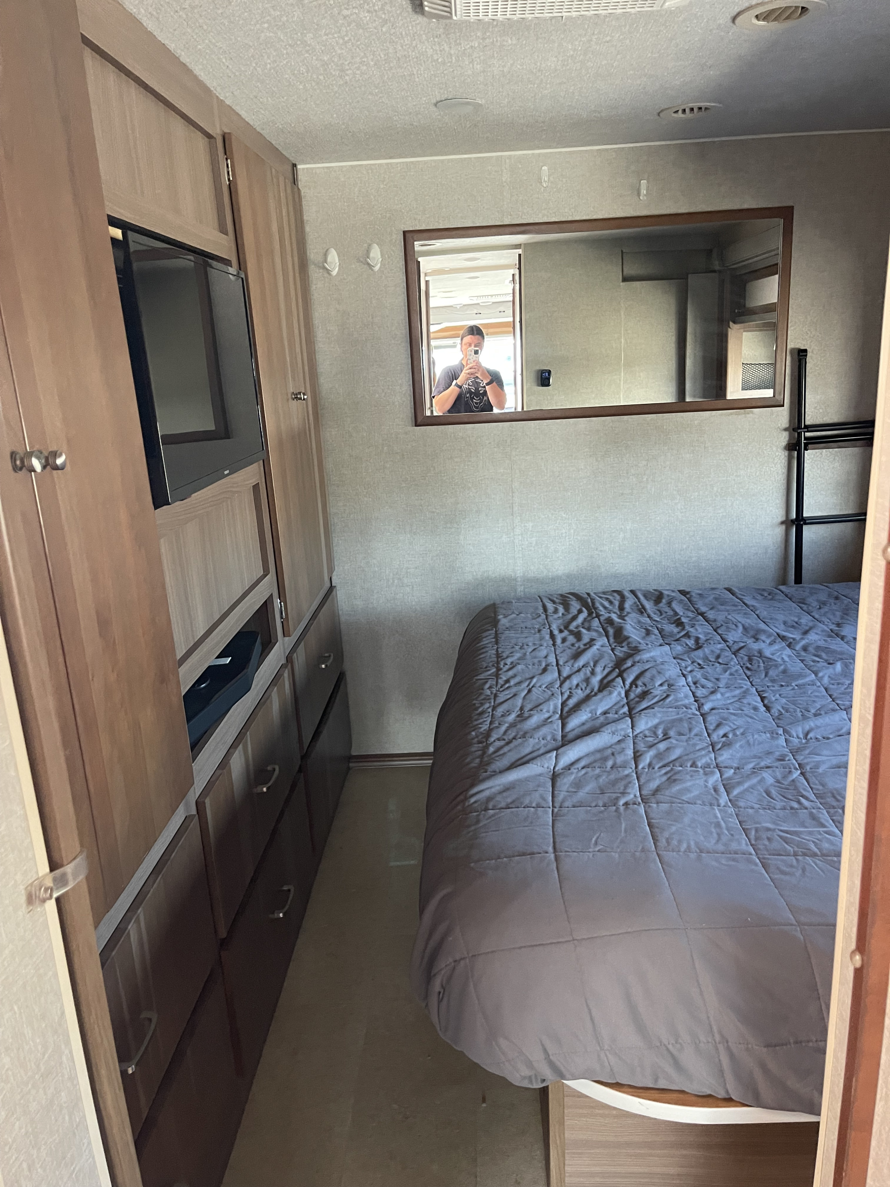 RV rental bedroom with cozy queen bed, TV, and built-in storage. Mirror reflects person and interior, ideal for travel.. Itasca Tribute 2015