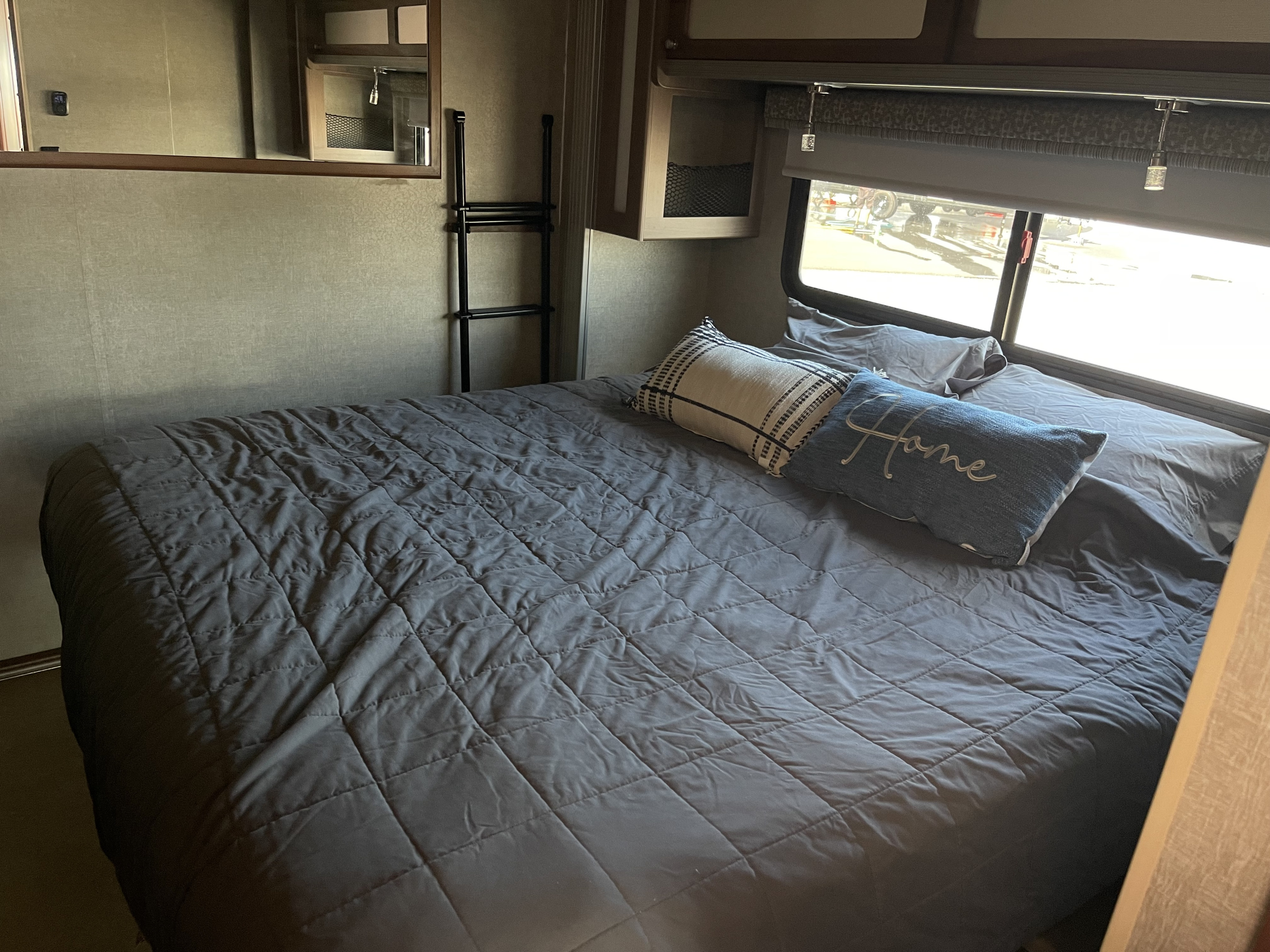 Cozy RV rental bedroom featuring a quilt-covered bed with "Home" pillow, a window with a view, and a ladder by the wall.. Itasca Tribute 2015
