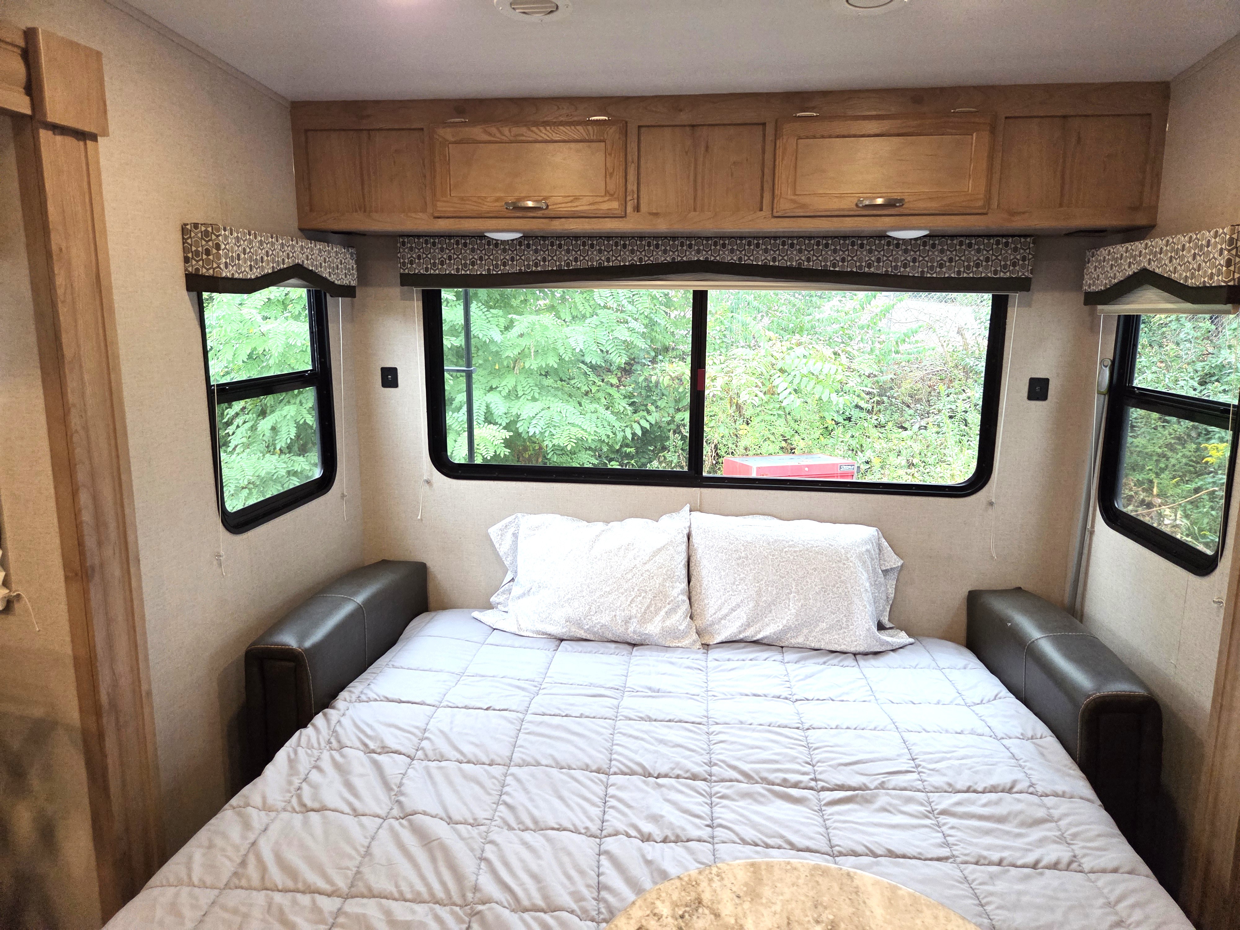 Cozy RV rental bedroom featuring a bed with light grey bedding, large windows viewing nature, and overhead storage cabinets.. Winnebago Minnie Winnie 2020