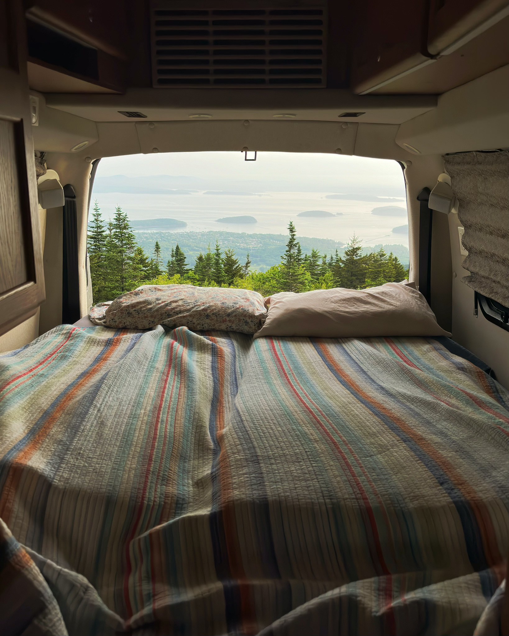 Cozy RV rental bed with a striped quilt, offering a scenic view of evergreen trees, islands, and ocean through the open back doors.. Roadtrek Ranger Rt 2012