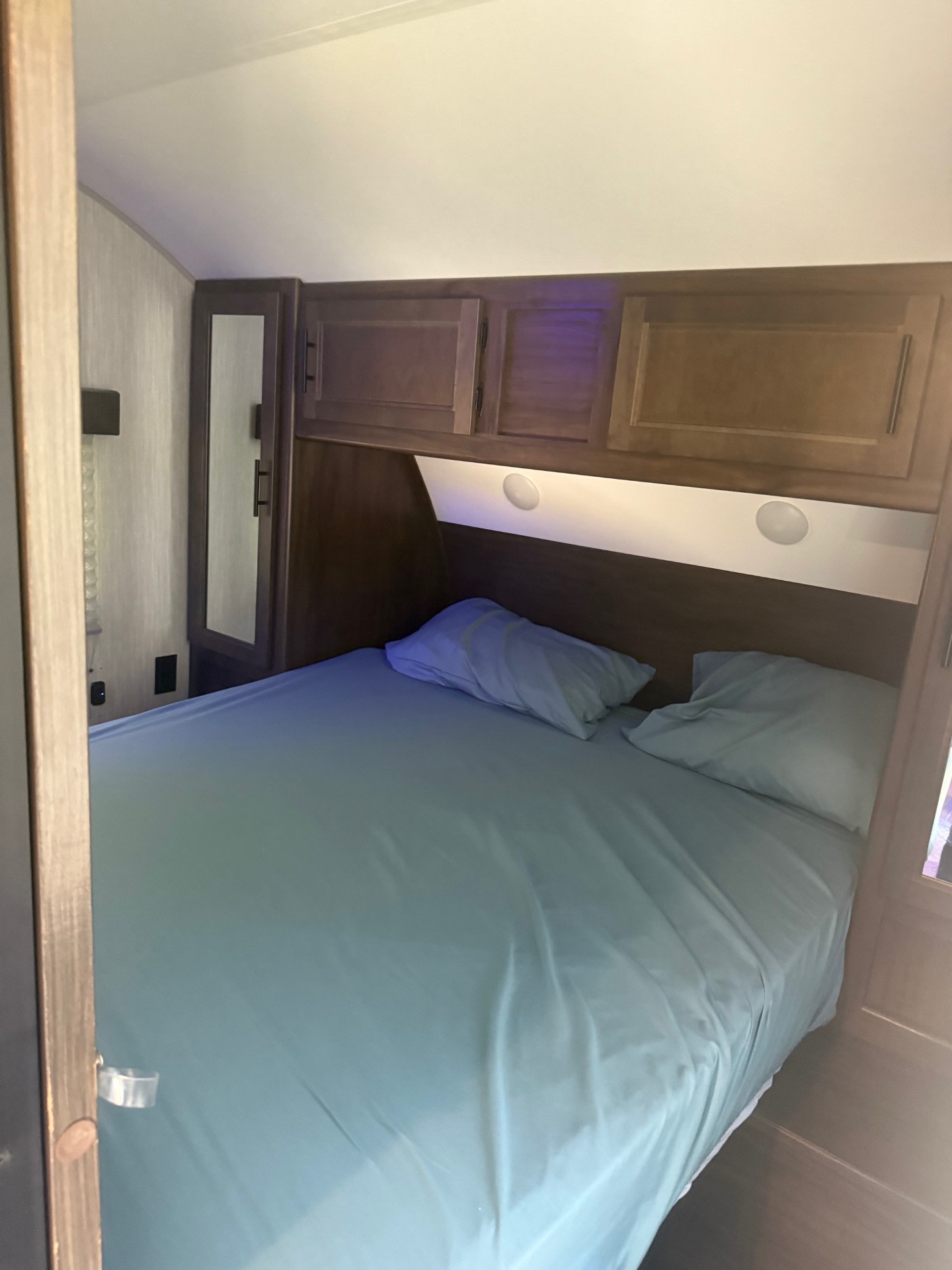 RV rental bedroom with a neatly made bed (blue sheets, pillows) and wood overhead cabinets, offering storage and comfort.. Forest River Cherokee Grey Wolf 2021