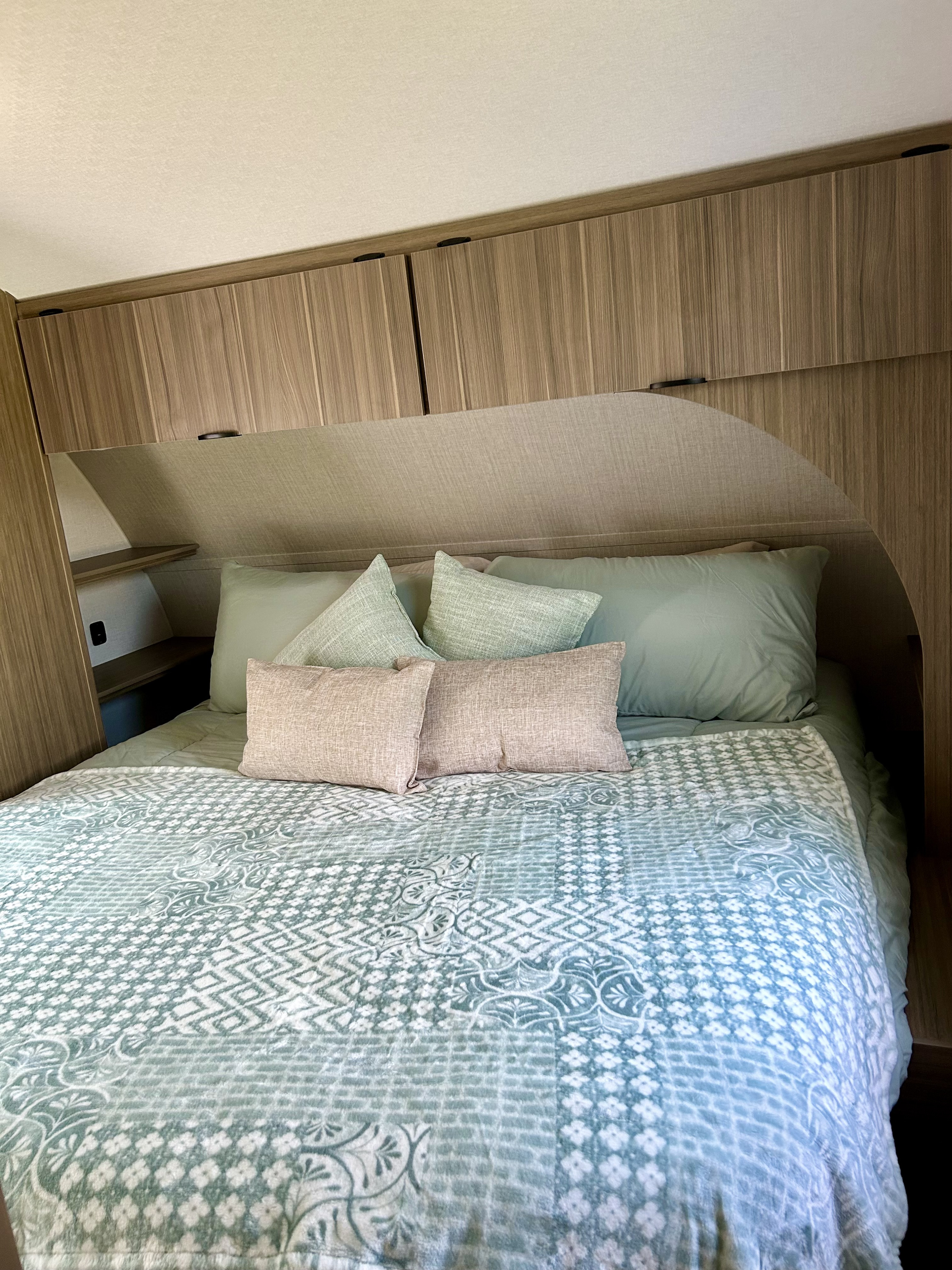 Cozy RV rental bedroom with soft green bedding, patterned duvet, and wood-grain overhead storage cabinets.. Forest River Wildwood X-Lite 2026