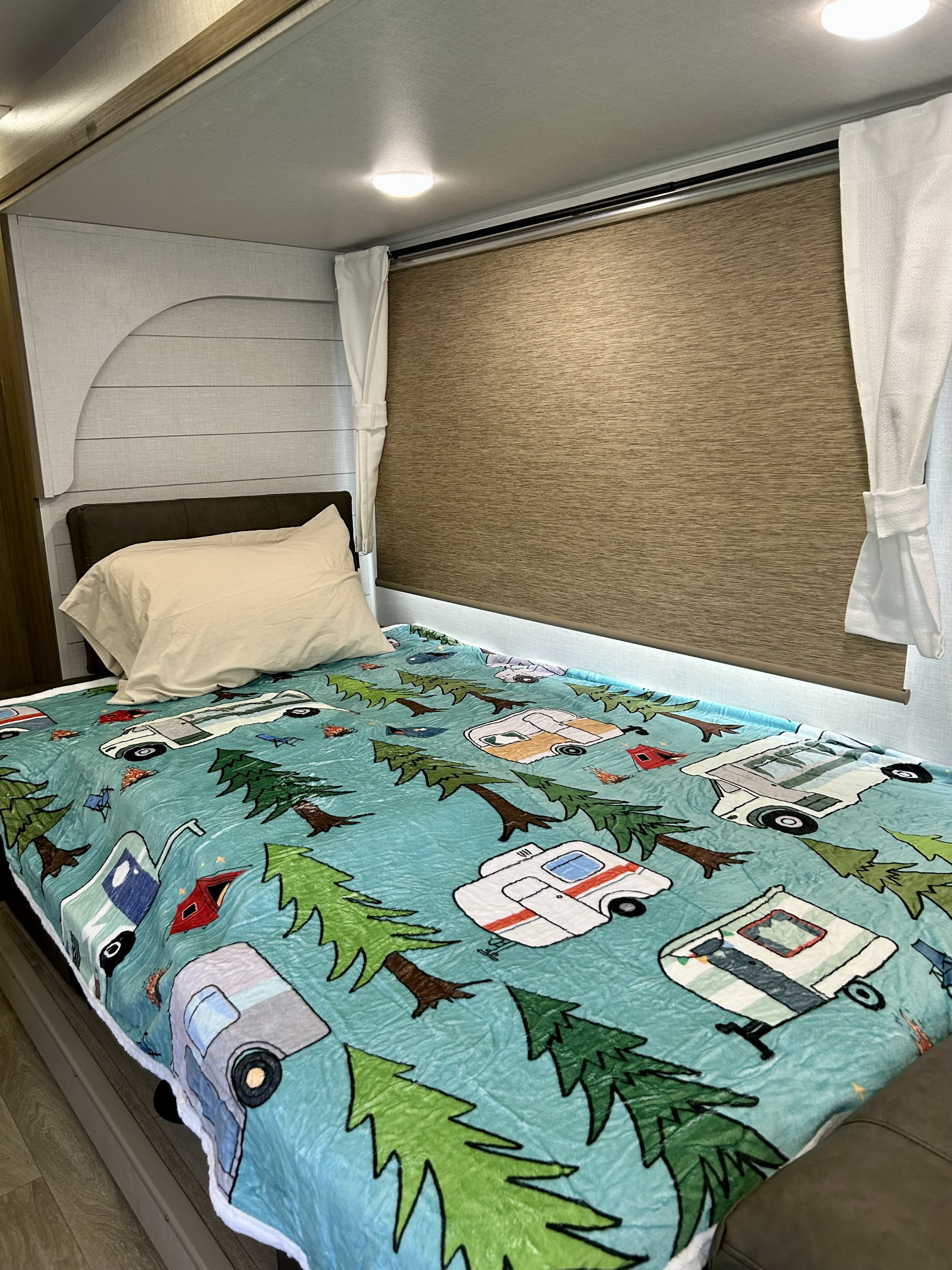 Cozy RV rental bed with a fun camping-themed blanket, pillow, and window blind. Perfect for a road trip adventure!. Forest River Wildwood X-Lite 2026