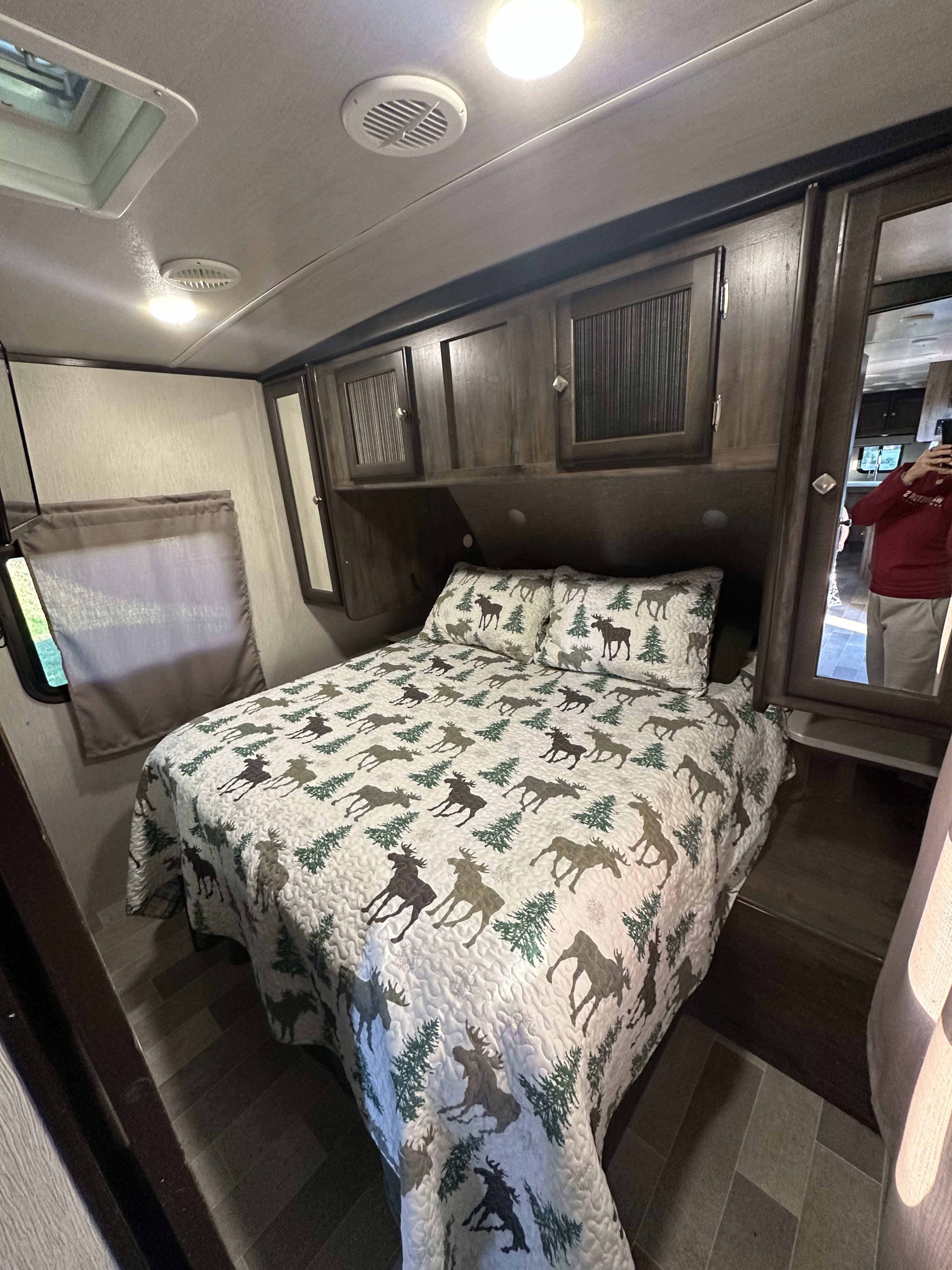 Cozy RV rental bedroom interior featuring a bed with a festive moose and pine tree quilt, overhead cabinets, and a window.. Heartland RVs Mallard 2020