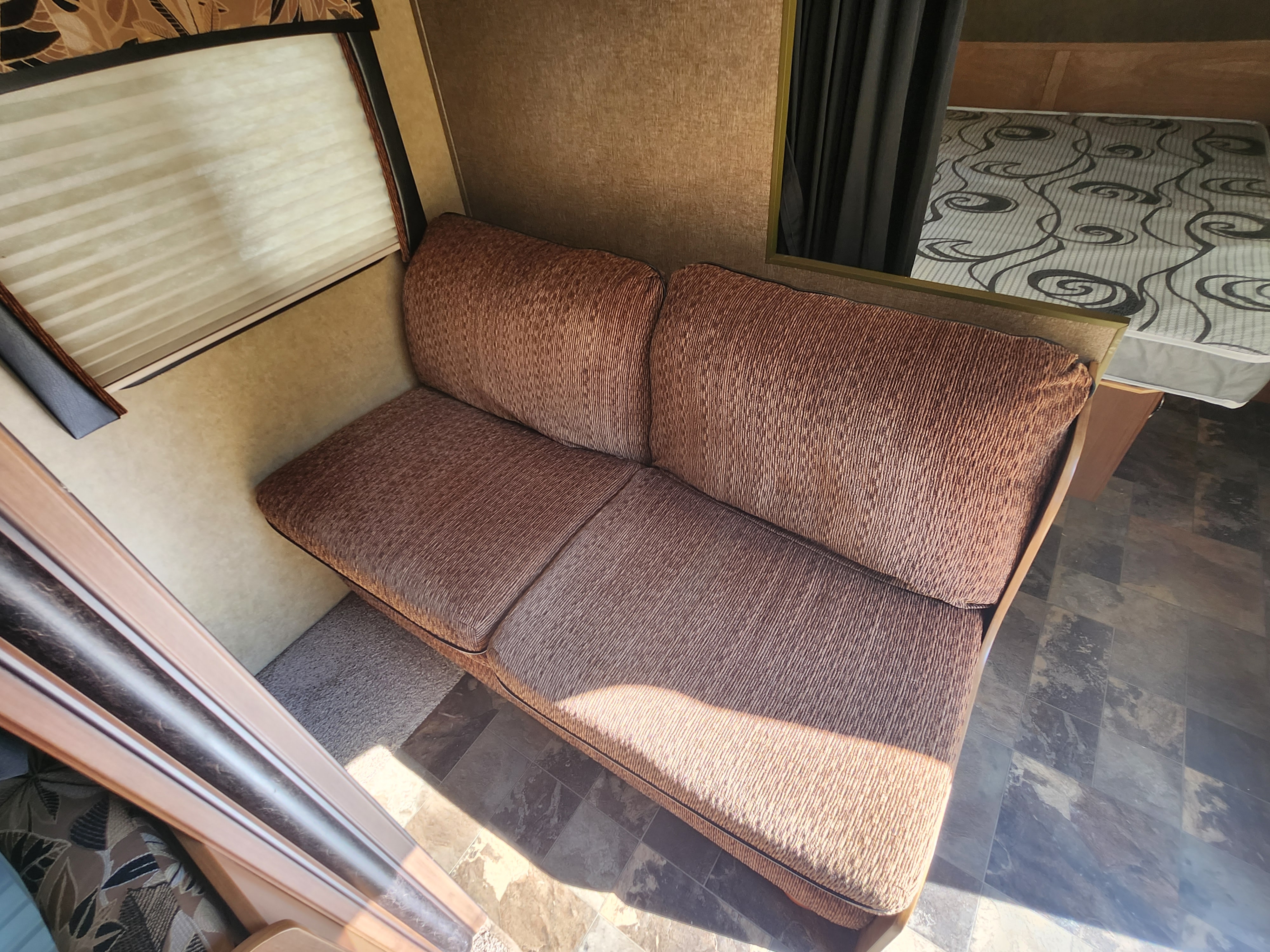 RV rental interior featuring a cozy brown sofa, window with blinds, and a glimpse of a patterned bed. Mixed flooring.. Starcraft Autumn Ridge 2013