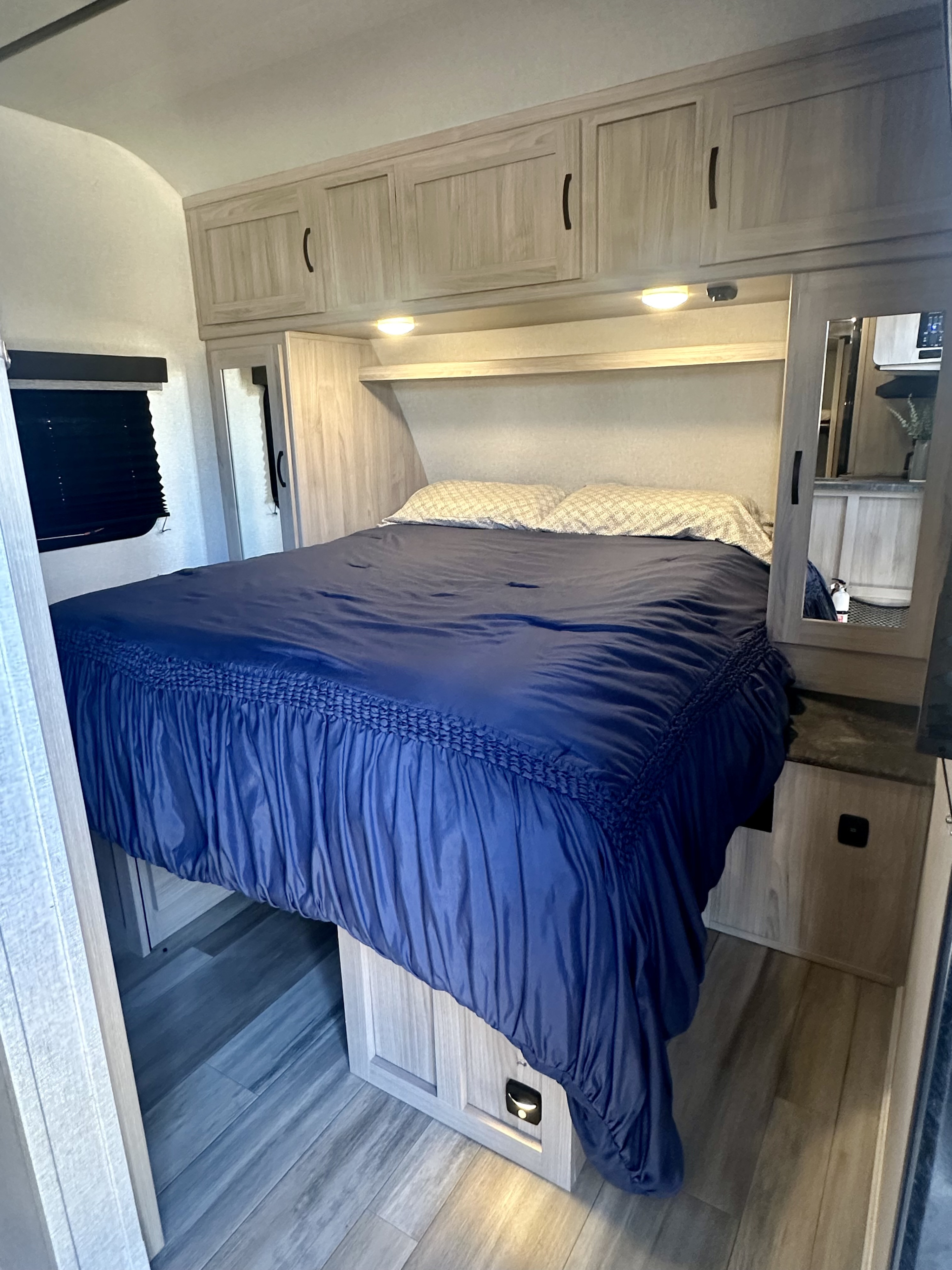 RV rental bedroom featuring a bed with dark blue bedding, overhead cabinets, a mirrored wardrobe, and light wood interior.. Coachmen 2963BH 2023