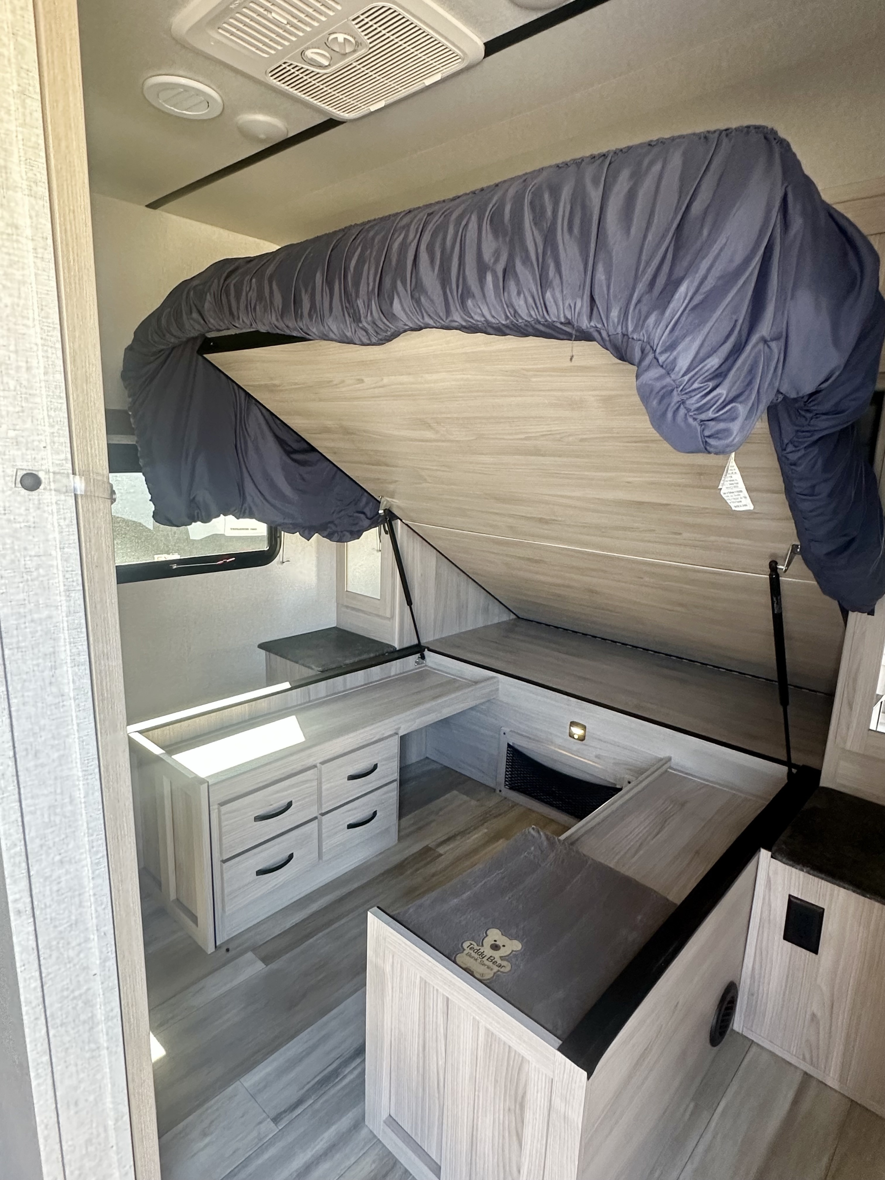 RV rental interior: space-saving lift-up bed reveals storage & desk area, with modern light-wood design and ceiling vents.. Coachmen 2963BH 2023