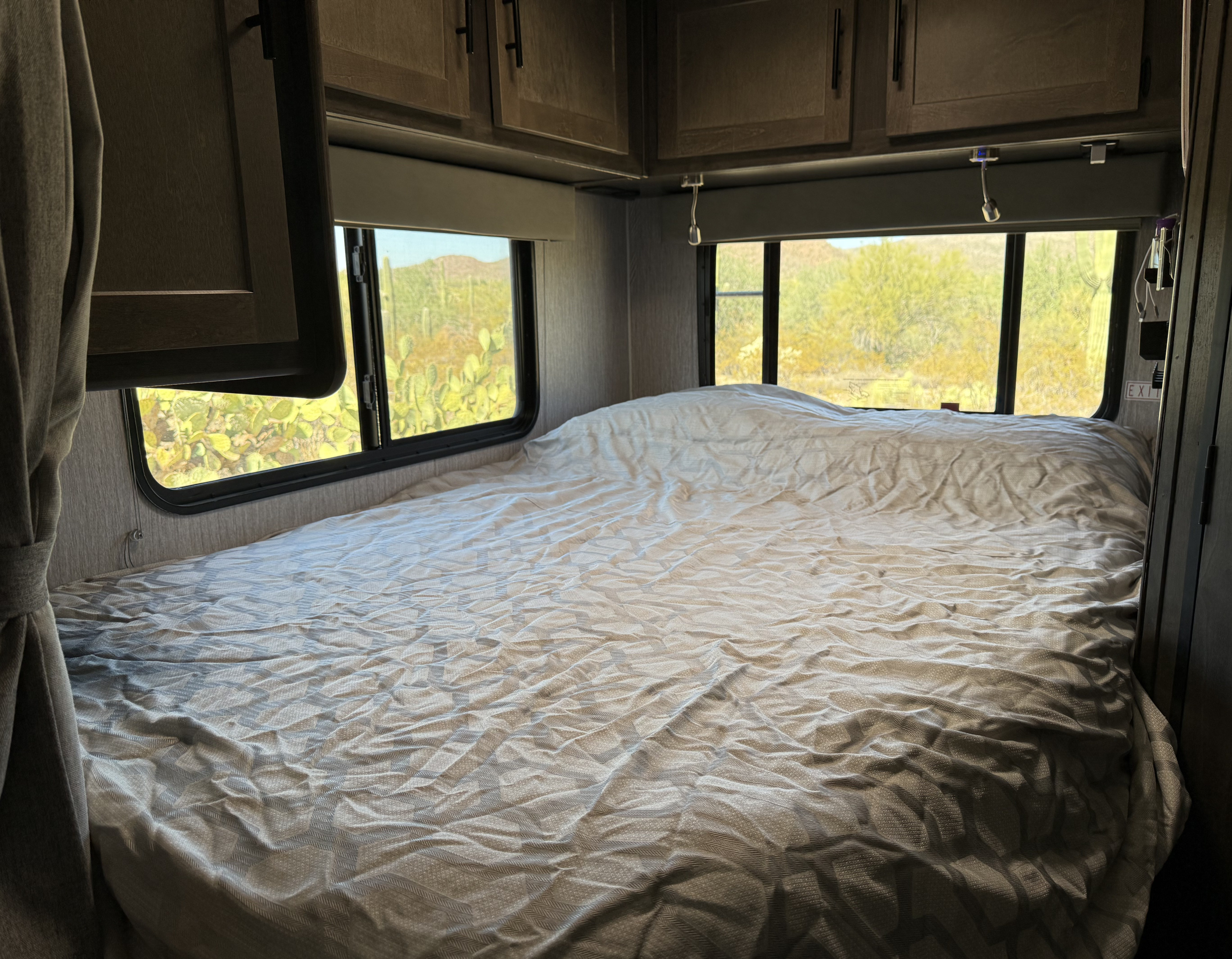 Relax in this RV rental bedroom featuring a bed with grey wrinkled bedding and a stunning desert view outside.. Forest River Coachmen Leprechaun 2022