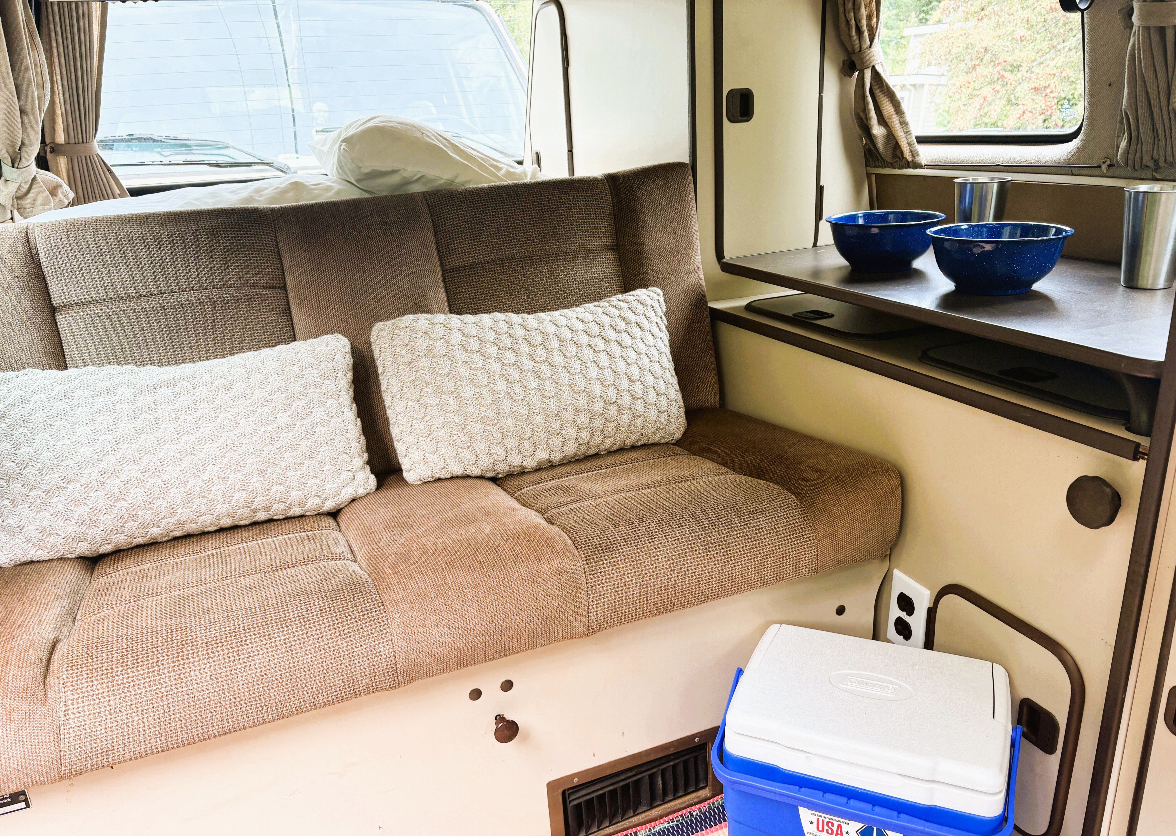 Cozy rv rental interior: A comfortable bench seat, pillows, dishes on a table, and a cooler. Ready for van life adventures!. Volkswagen Westfalia 1985