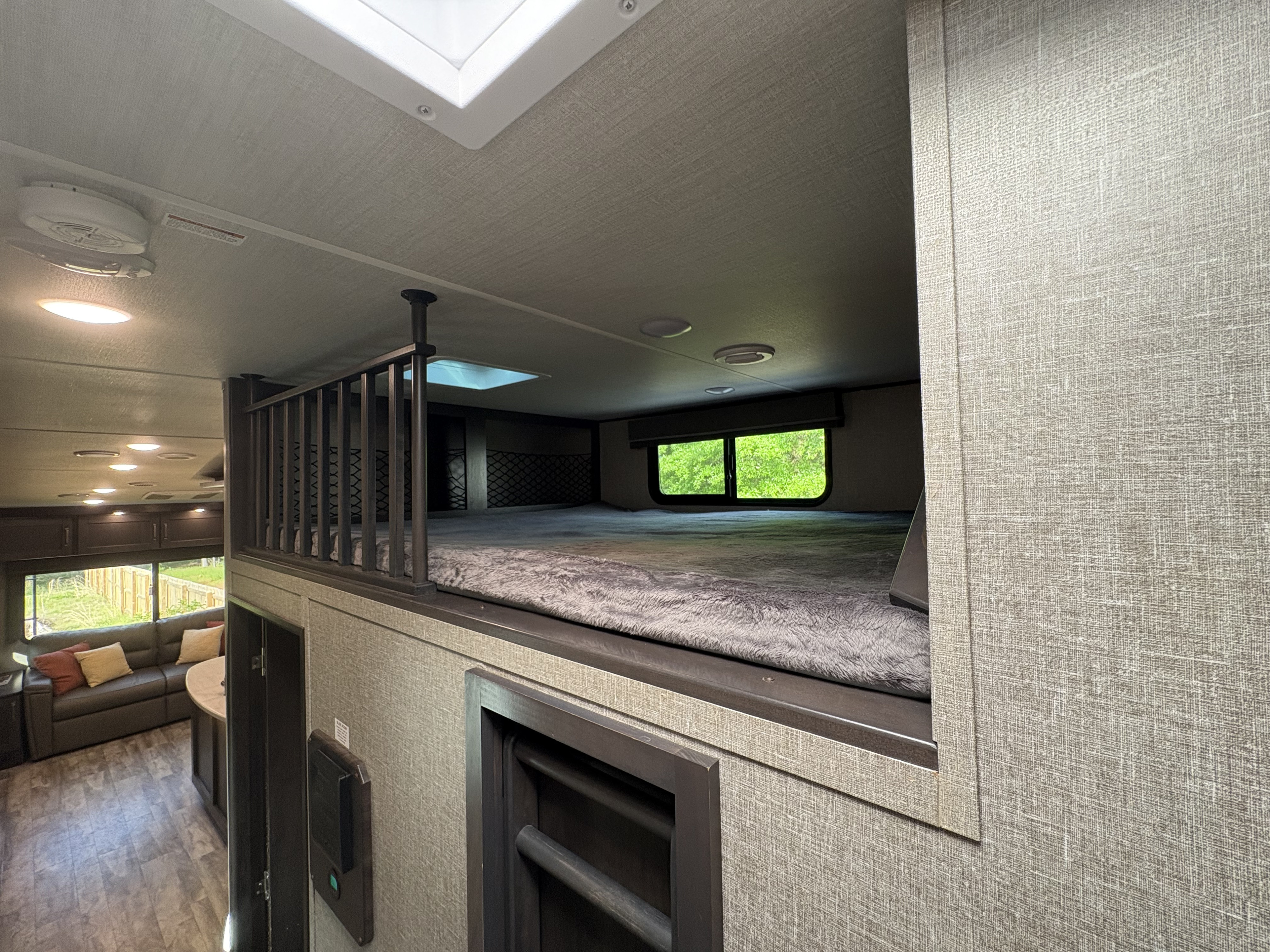 Inside an RV rental: A spacious loft bunk bed with a railing, skylight, and window overlooks a living area with a sofa.. Grand Design Reflection 2023