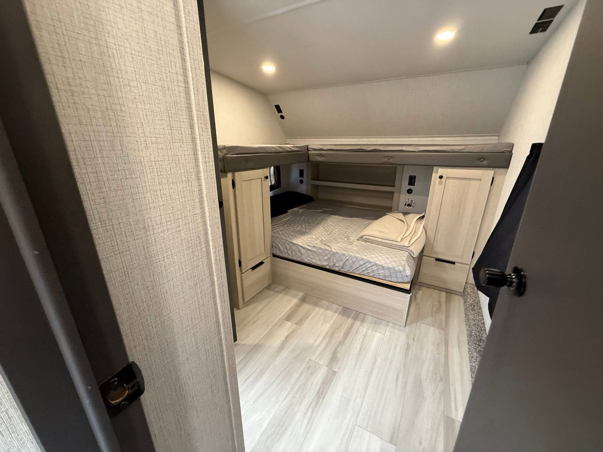 RV rental bedroom: light wood interior, bed, and upper bunk beds, illuminated by recessed lighting, seen from a doorway.. Forest River Cardinal 2025