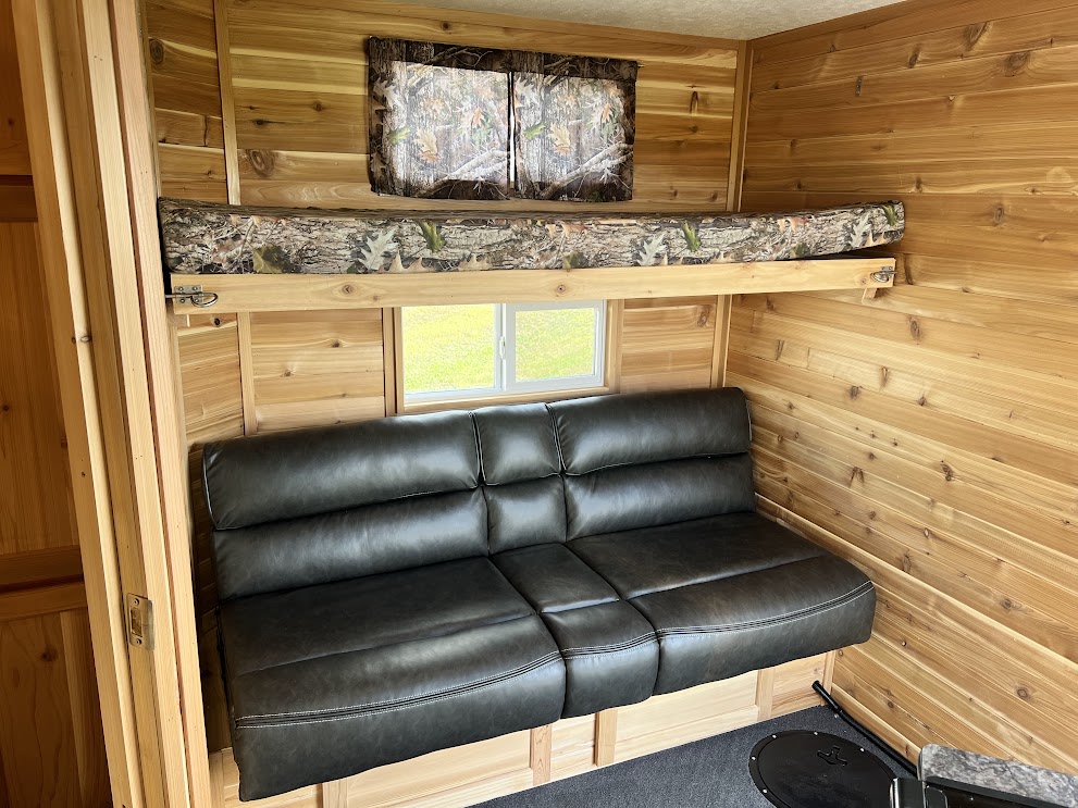 This RV rental interior features natural wood walls, a dark grey sofa, camouflage bunk bed, and window with outdoor view.. Ice Castle Limited Deluxe 2023