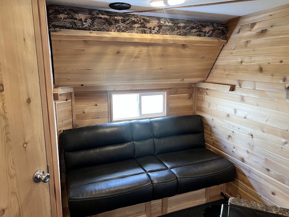This RV rental interior features natural wood walls, a comfy black sofa, and an overhead bunk with camouflage bedding.. Ice Castle Limited Deluxe 2023