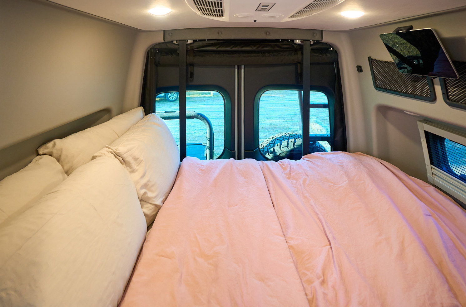 Comfortable bed inside an RV rental, featuring white pillows, a pink duvet, and a view through the rear van windows.. Winnebago Revel 2026