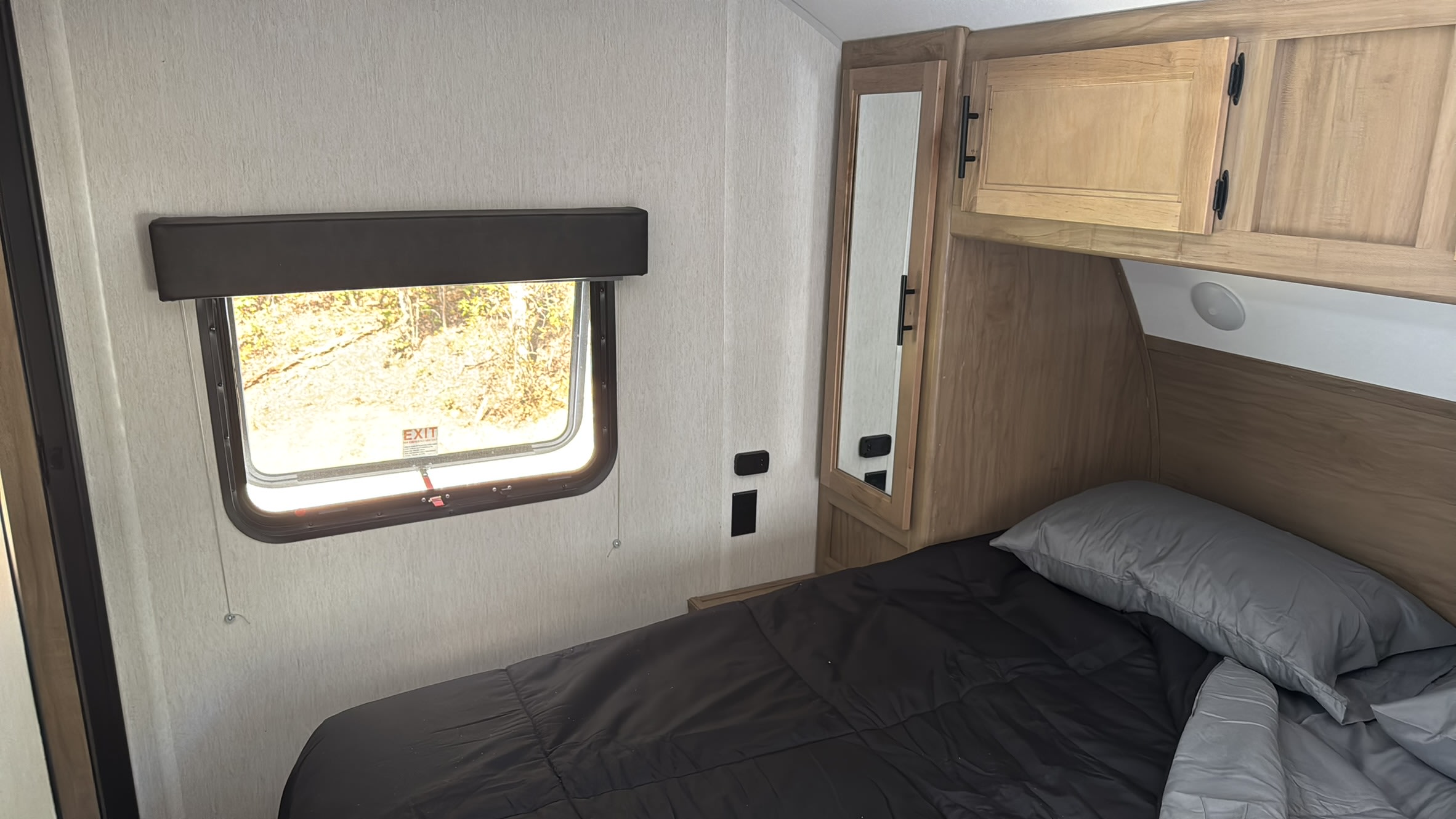 Inside an RV rental bedroom: a comfy bed with black comforter, window with an "EXIT" sign, and wooden storage.. Forest River Cherokee Grey Wolf 2023