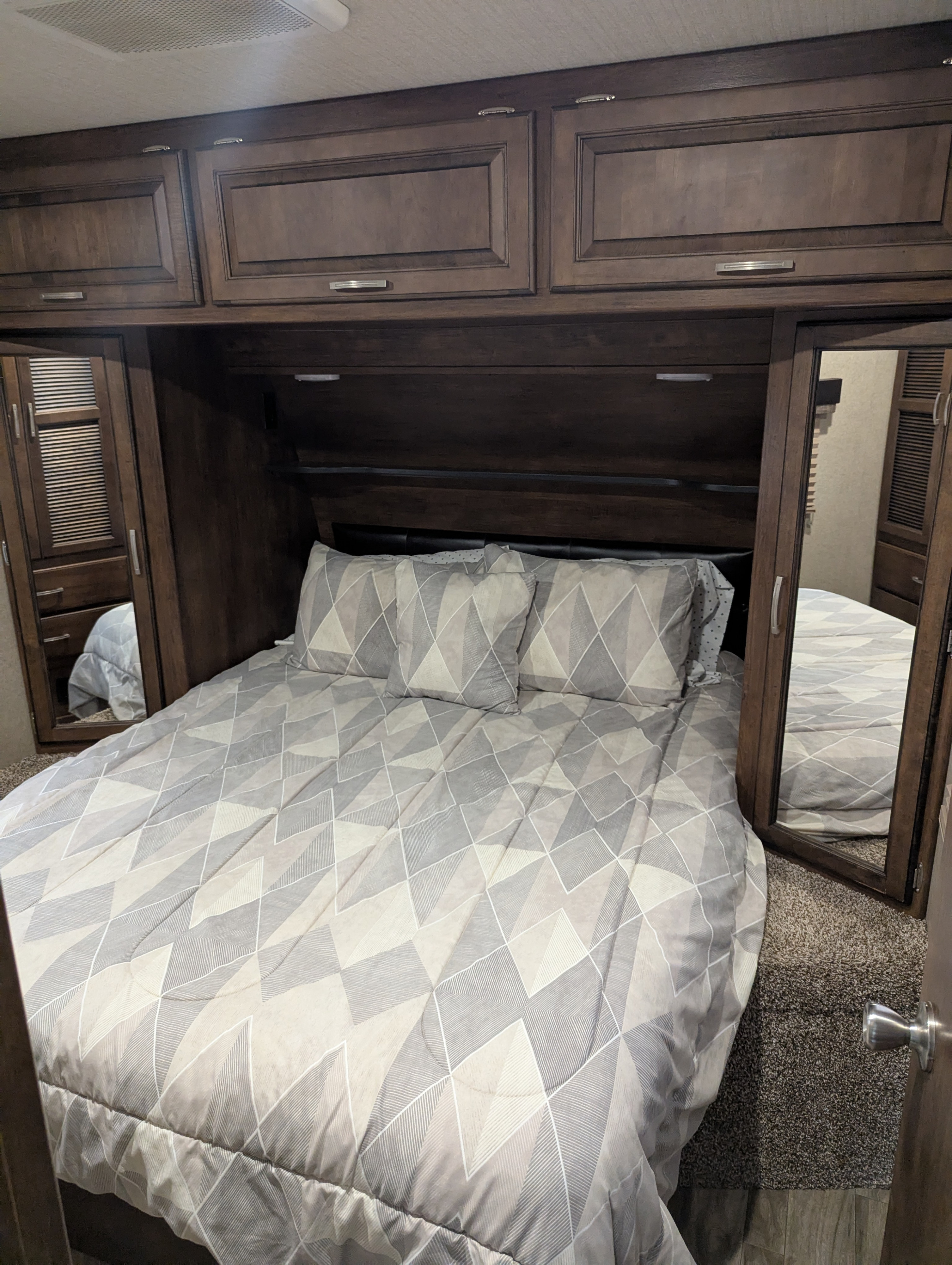 Inside an RV rental, a bedroom with a bed, patterned duvet and pillows, mirrored wardrobes, and dark wood overhead cabinets.. Grand Design Reflection 2019
