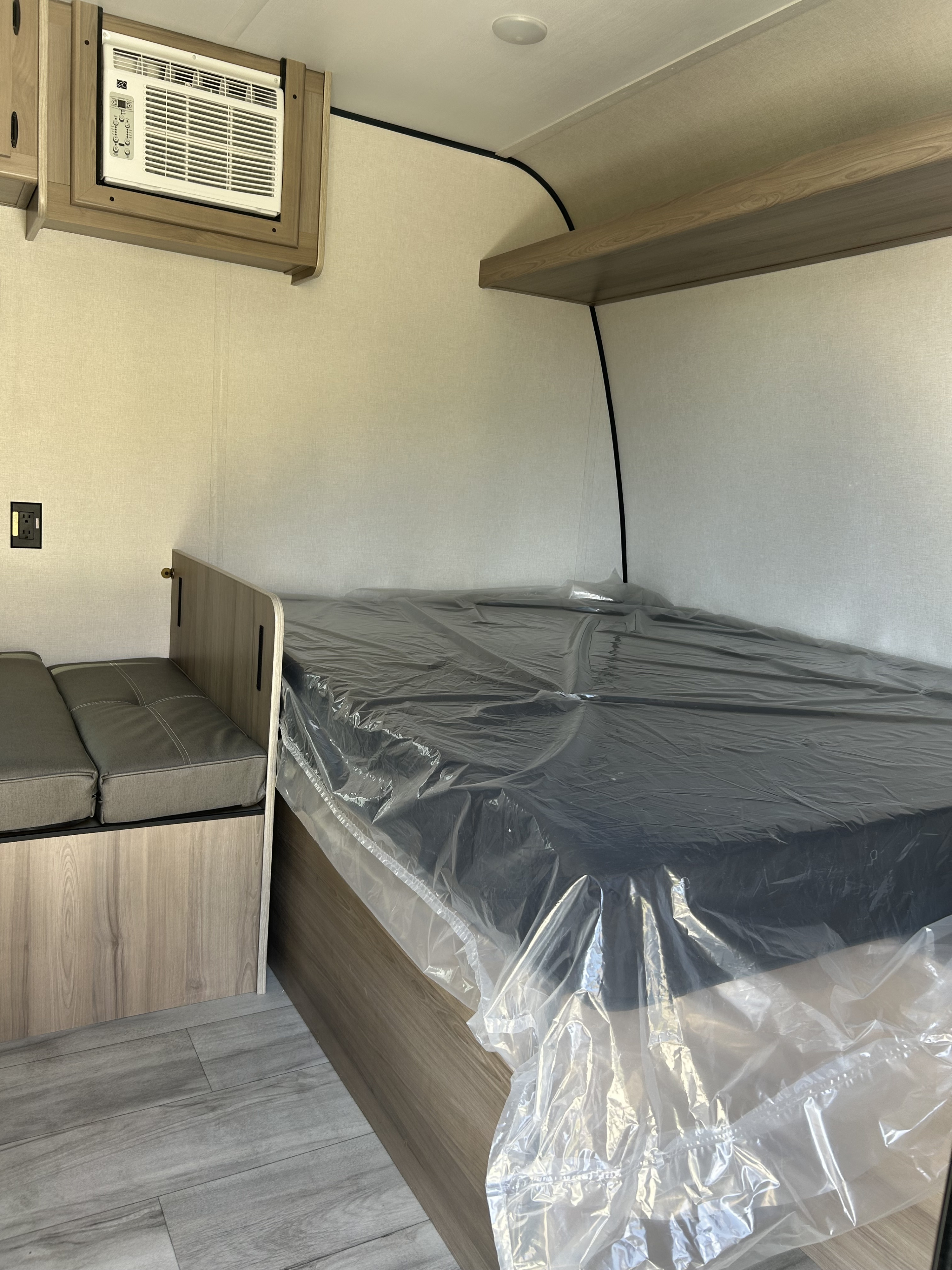 RV rental interior: new bed (plastic-wrapped), side seating, AC unit, and wall shelf in a modern travel trailer.. Coleman Lantern 17B 2025