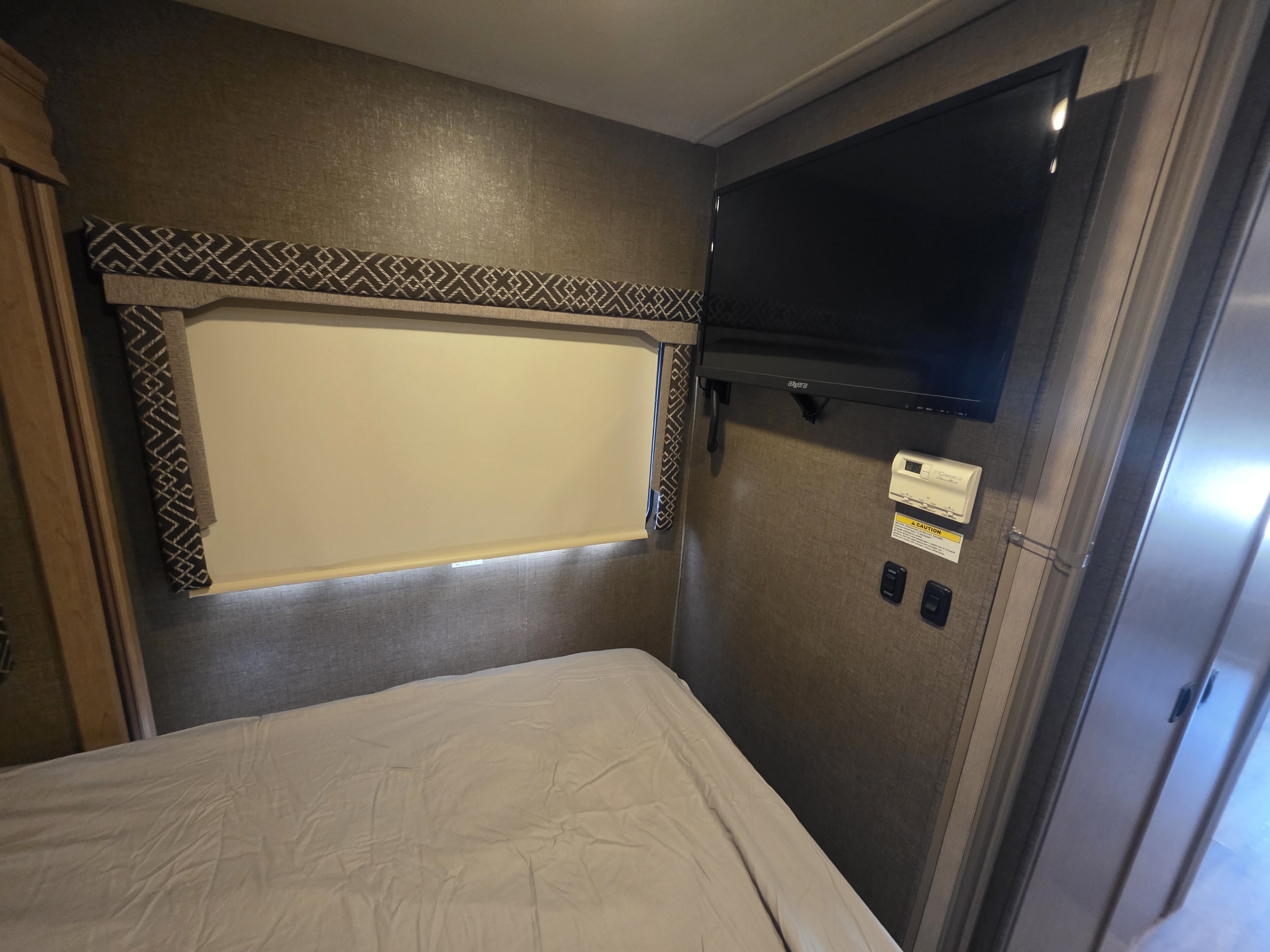 RV rental bedroom with a neatly made bed, window with patterned valance, wall-mounted TV, and climate controls.. Thor Four Winds 2018