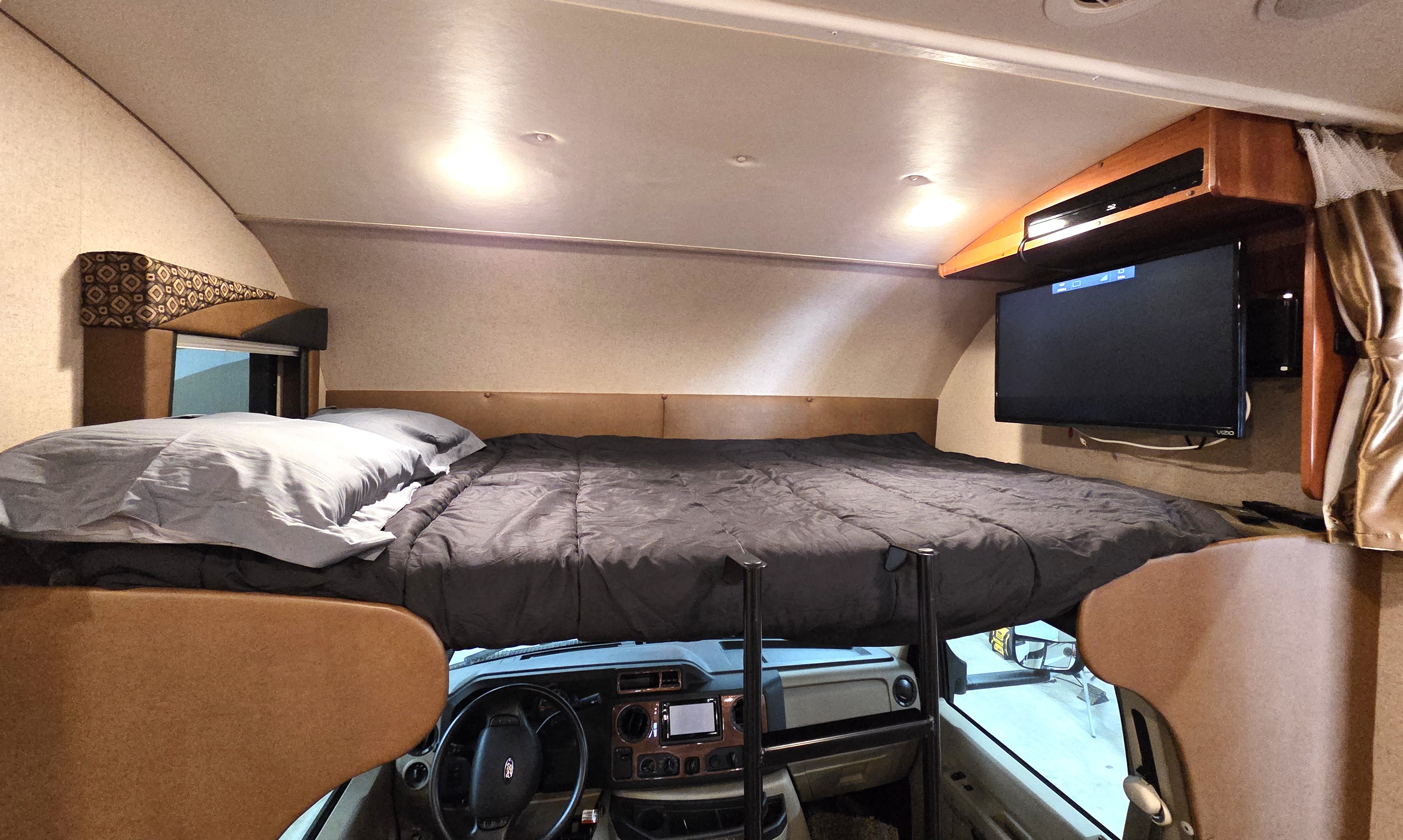Inside an RV rental: a cozy cab-over bed overlooking the driver's cockpit, complete with a TV for comfort.. Jayco Greyhawk 2015