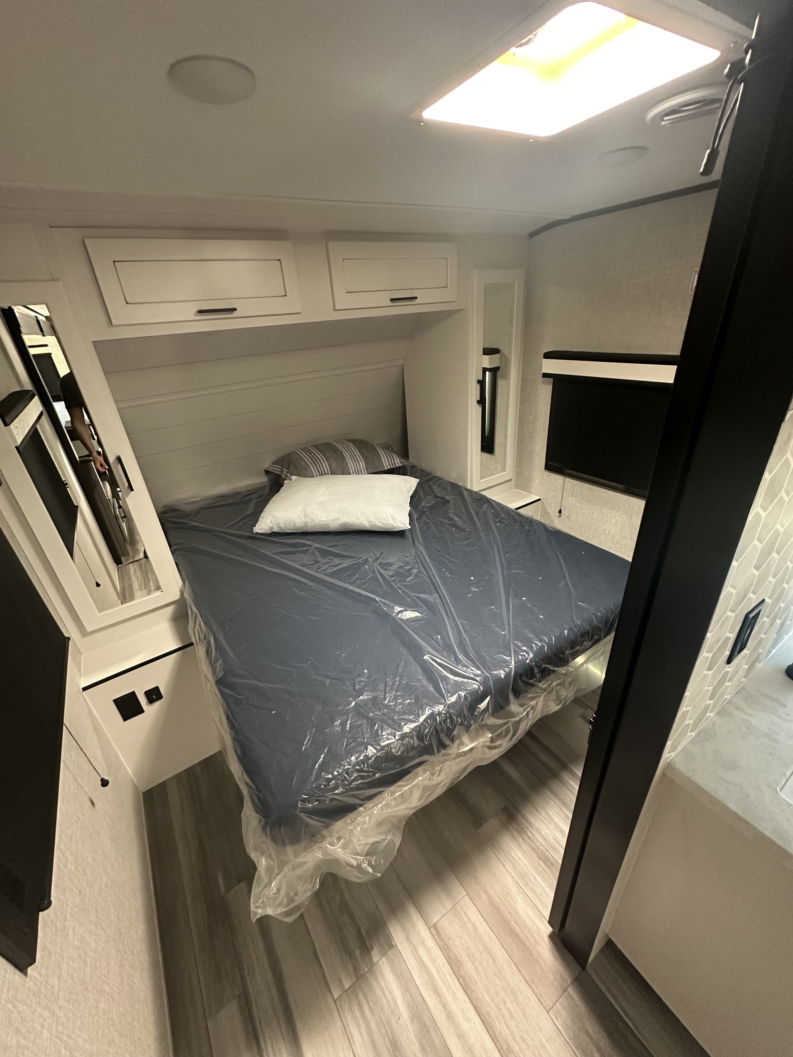 RV rental bedroom with bed in plastic, pillows, overhead cabinets, mirrors, and a bright skylight. Ready for travel.. Jayco Jay Feather 2024