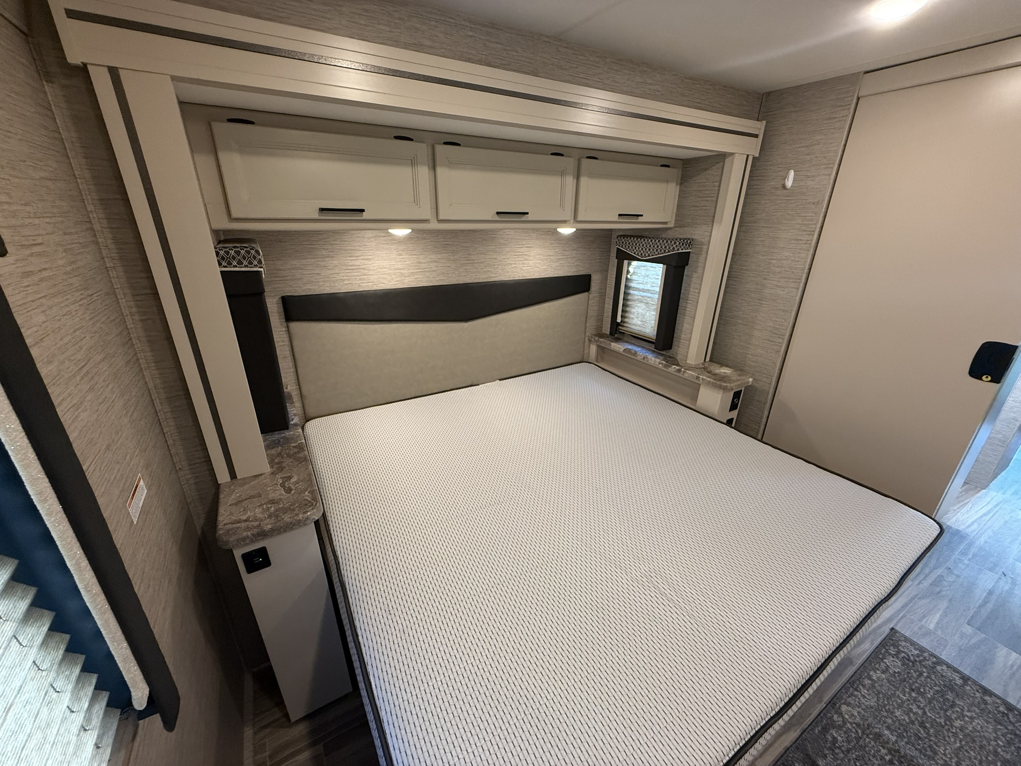 This RV rental bedroom features a queen bed, overhead storage with lights, and a small window, offering a comfortable interior.. Thor A.C.E. 2022