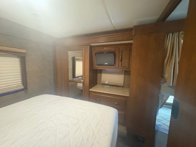 Cozy rv rental bedroom: main bed with white quilt, TV, storage, window, and a glimpse of an adjacent bunk bed through an opening.. A.C.E. A.C.E. Motorhome 2015