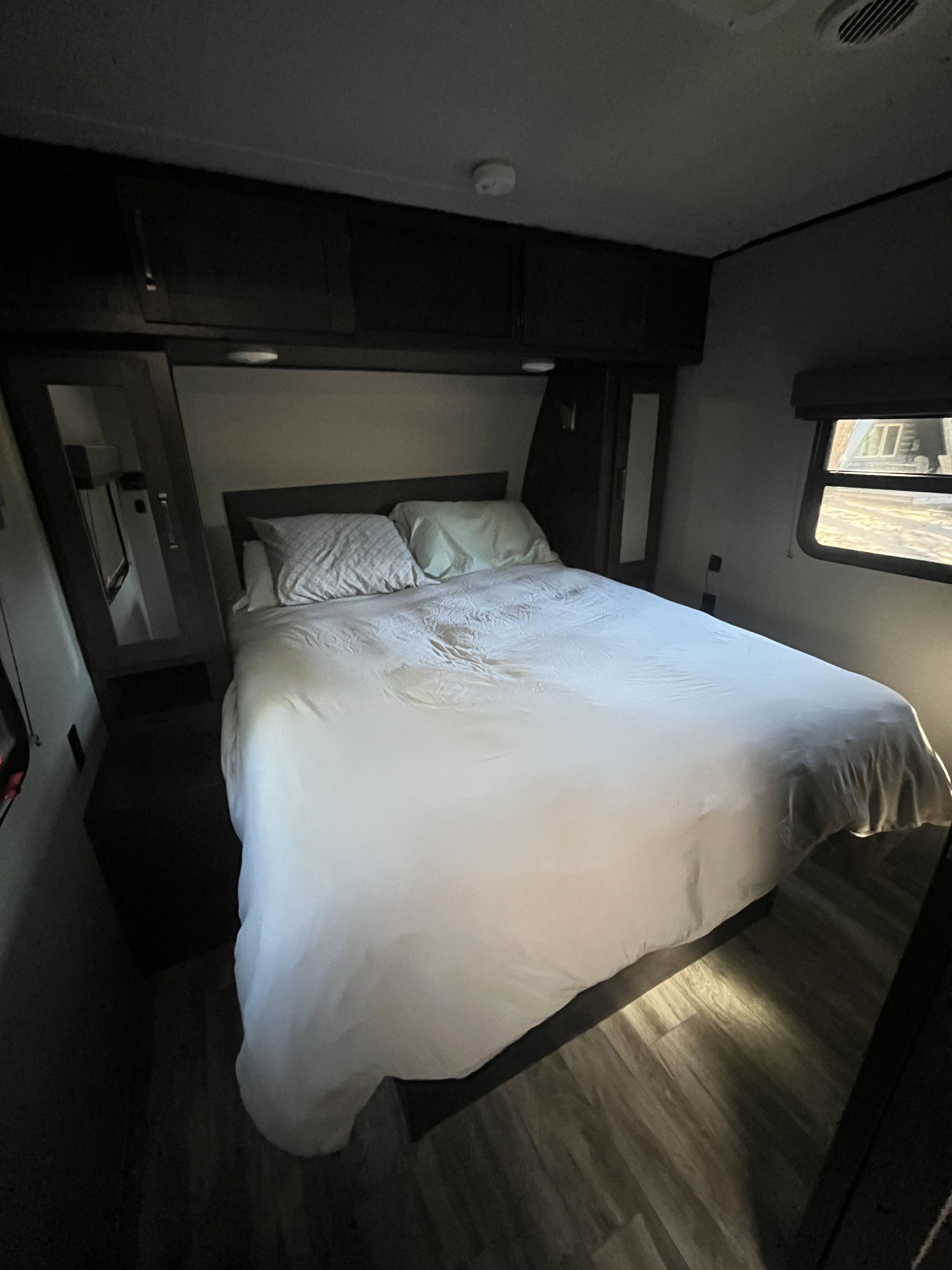 Dimly lit RV rental bedroom featuring a queen bed, overhead storage, wood-look flooring, and a window.. Grand Design Transcend 2020