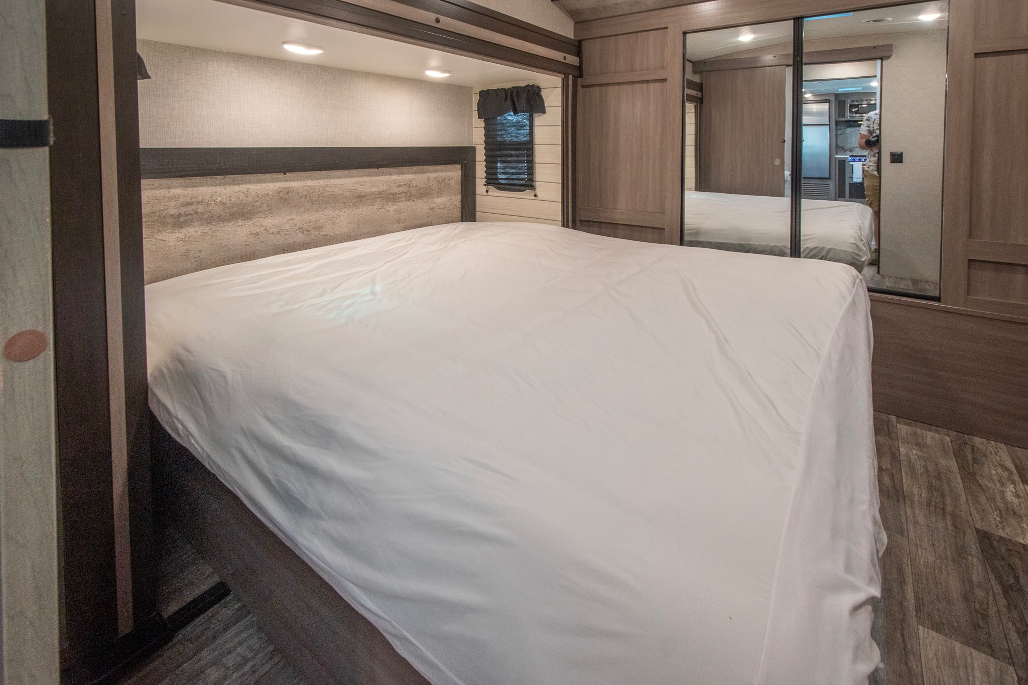 Spacious RV rental bedroom featuring a white bed, wood-textured headboard, window, and mirrored closet reflecting interior space.. Crossroads RV Sunset Trail 2022