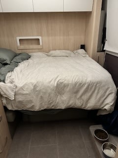 Compact RV rental bedroom showing a bed with light bedding, light wood walls, overhead cabinets, and pet bowls on a tiled floor.. Leisure Travel Vans Unity 2023