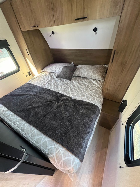 Modern rv rental bedroom: bed with patterned sheets, overhead cabinets, and sconce lights. Light wood interior and small windows.. Cherokee Grey Wolf 2025