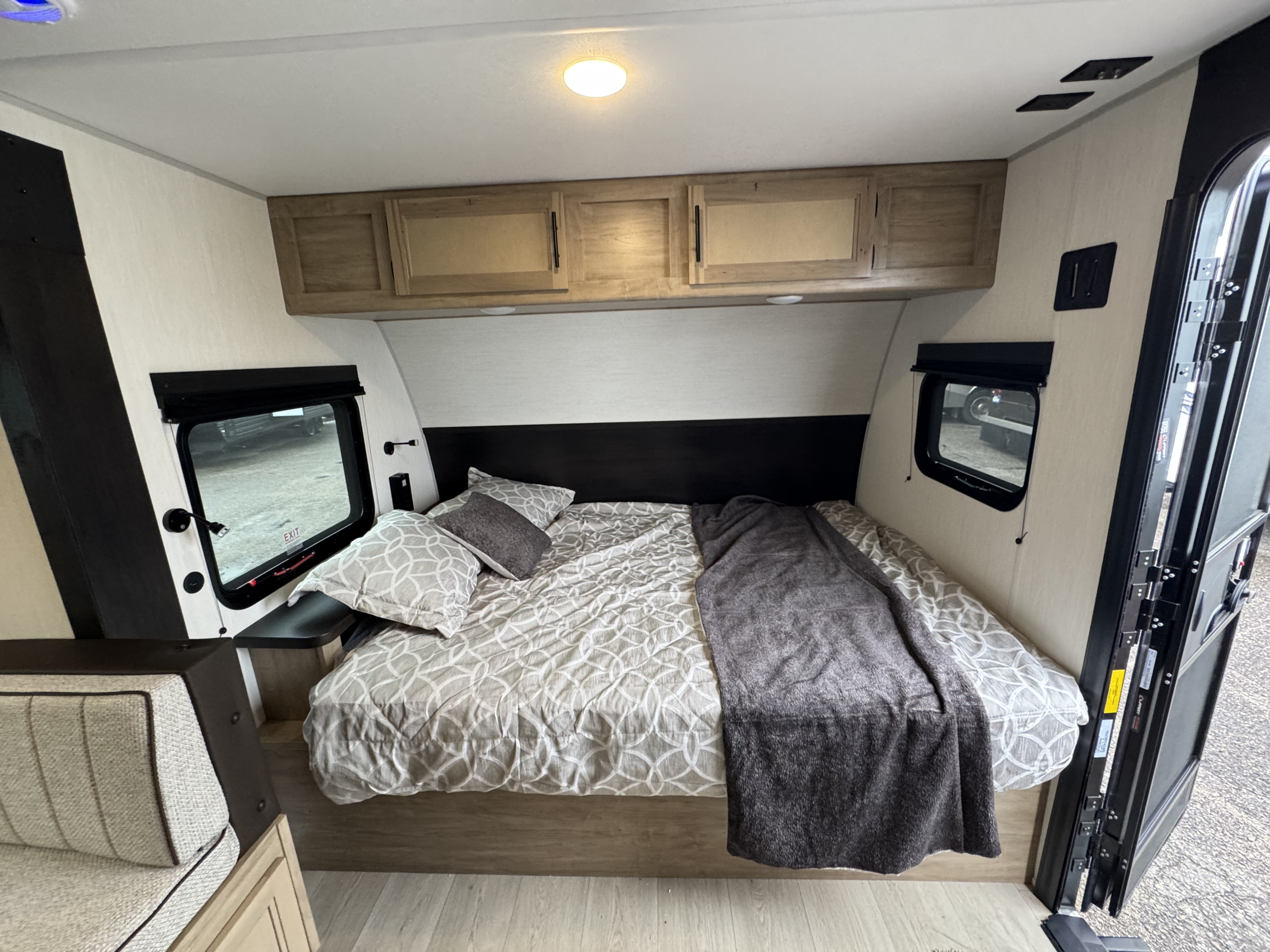 A warm and inviting RV rental bedroom with a patterned bed, dark wood headboard, overhead cabinets, and side windows.. Cherokee WolfPup 2025