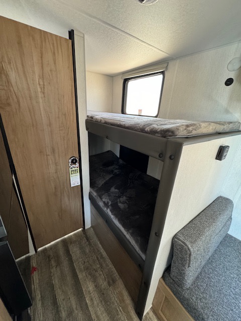RV rental bunk beds. A compact sleeping area featuring patterned mattresses, a window, wood-look floor, and a small grey bench.. Cherokee WolfPup 2025