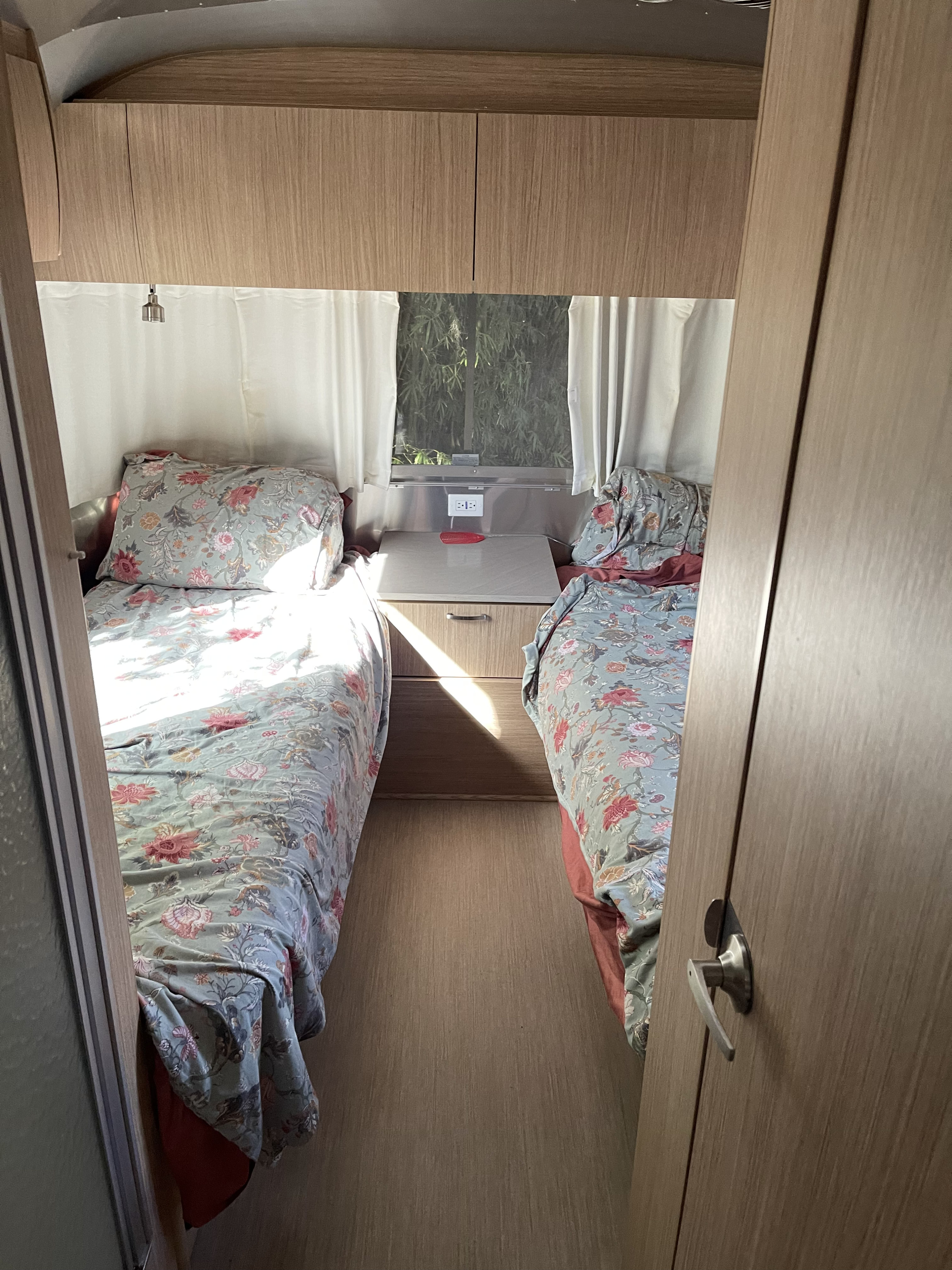 Bright RV rental bedroom: two beds with floral bedding, a central nightstand, overhead cabinets, and sunlight.. Airstream Flying Cloud 2019