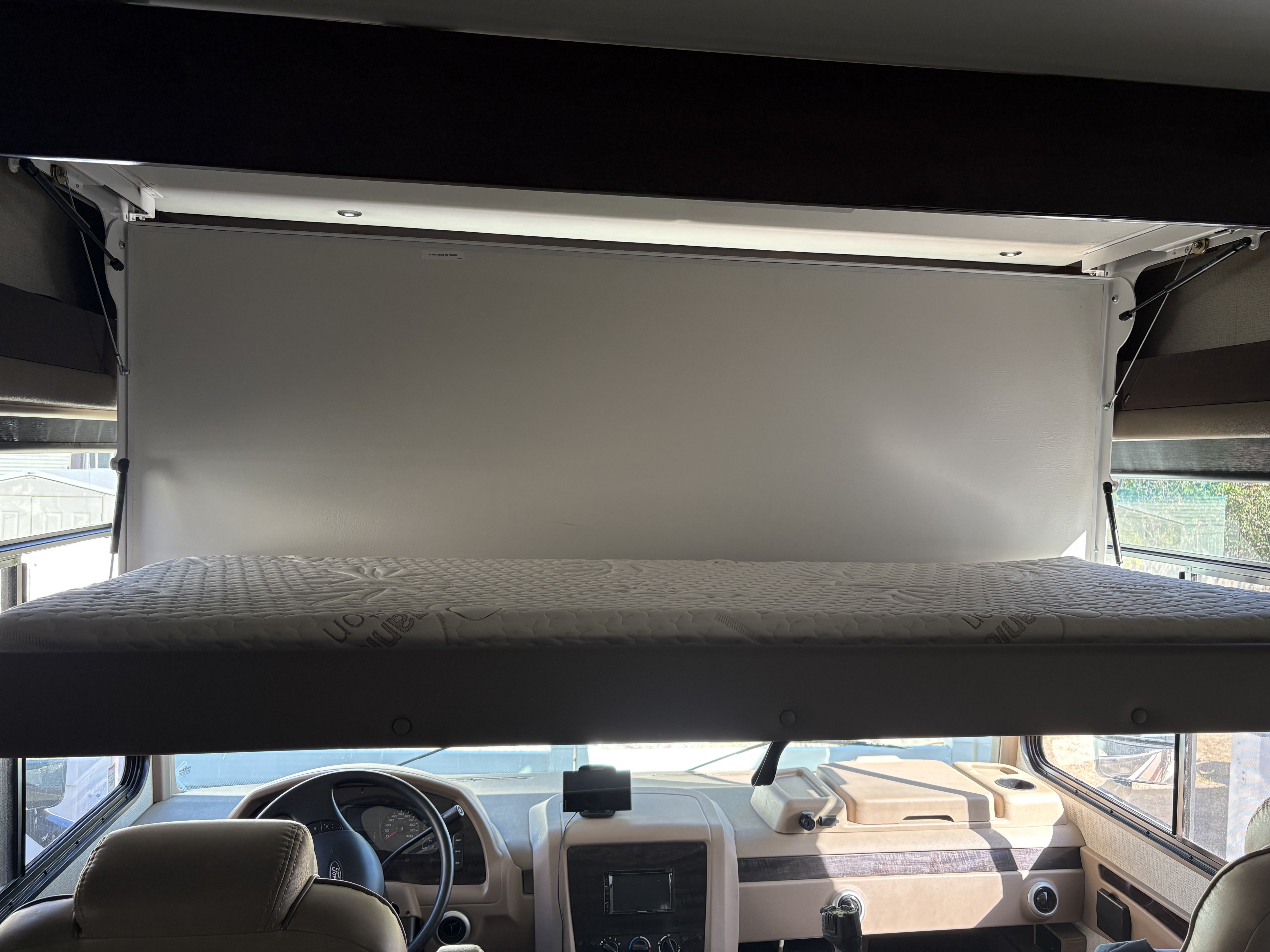 Overhead bunk bed lowered over the driver's cabin in an RV rental, showing a white mattress and dashboard view.. Georgetown XL 2019