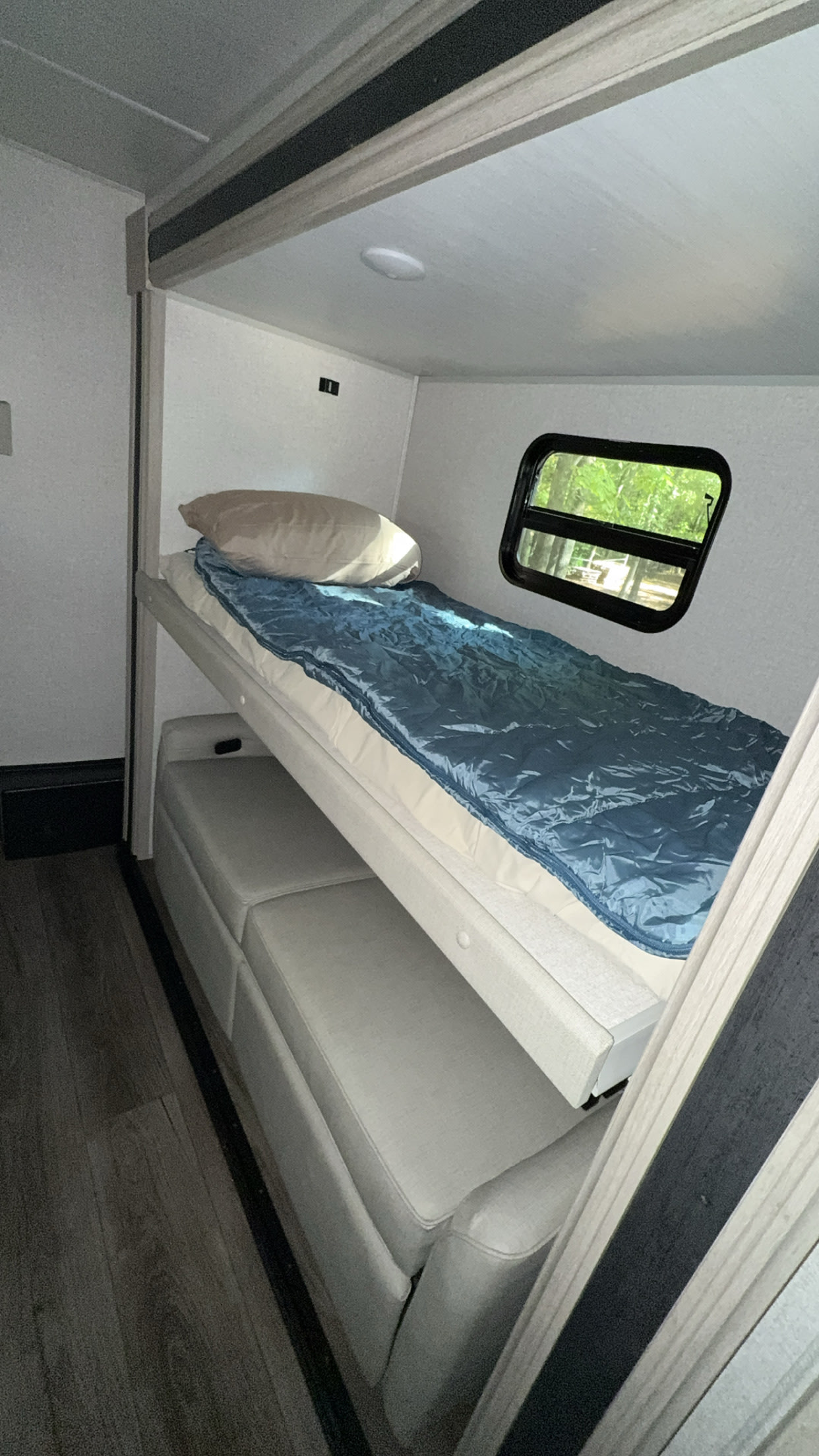 RV rental interior: cozy bunk bed setup. Top bunk with blue sleeping bag, bottom converts from sofa. Window shows green trees.. East to West Alta 2023