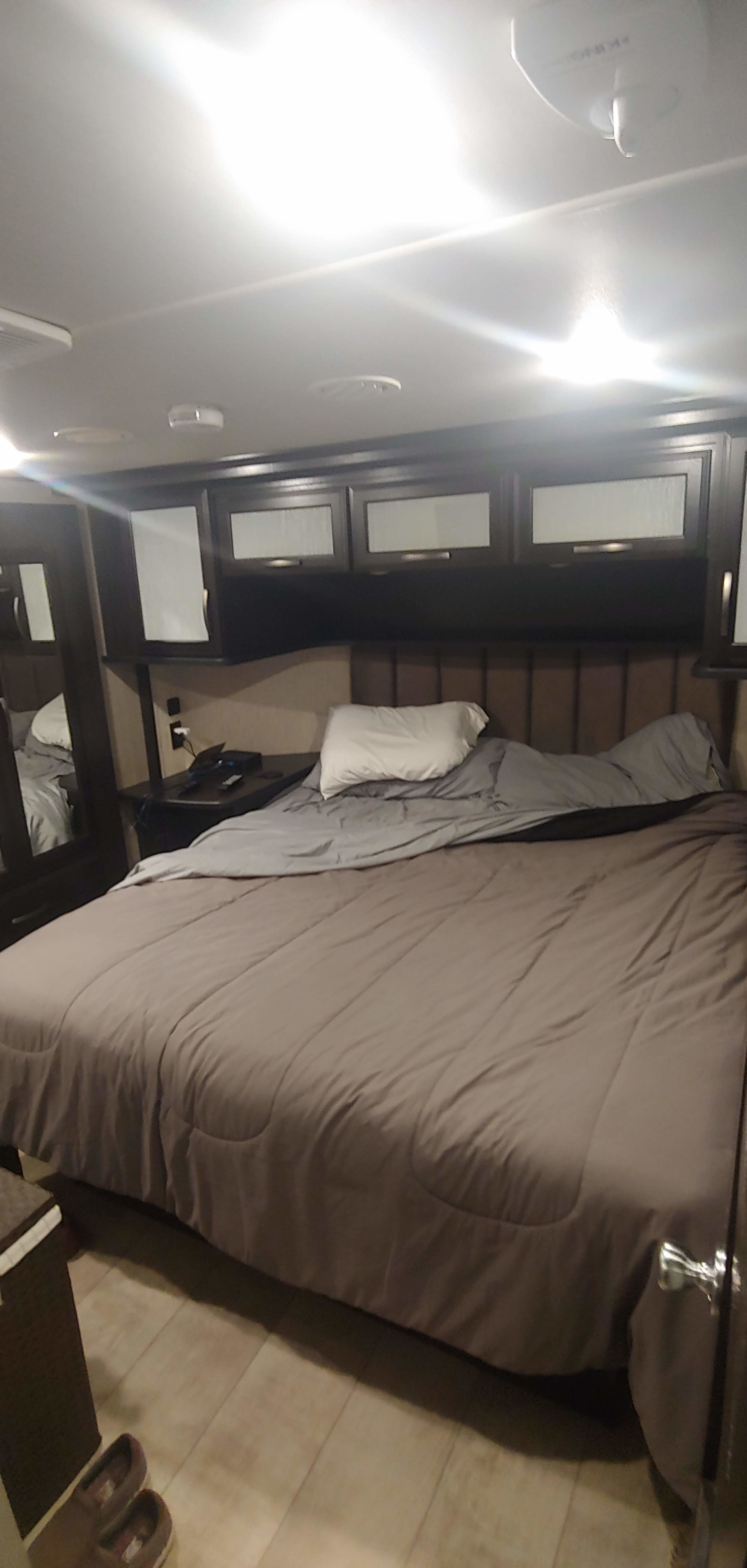 This RV rental bedroom offers a comfortable bed with grey bedding, overhead storage, and functional nightstand space.. Grand Design Momentum 2018