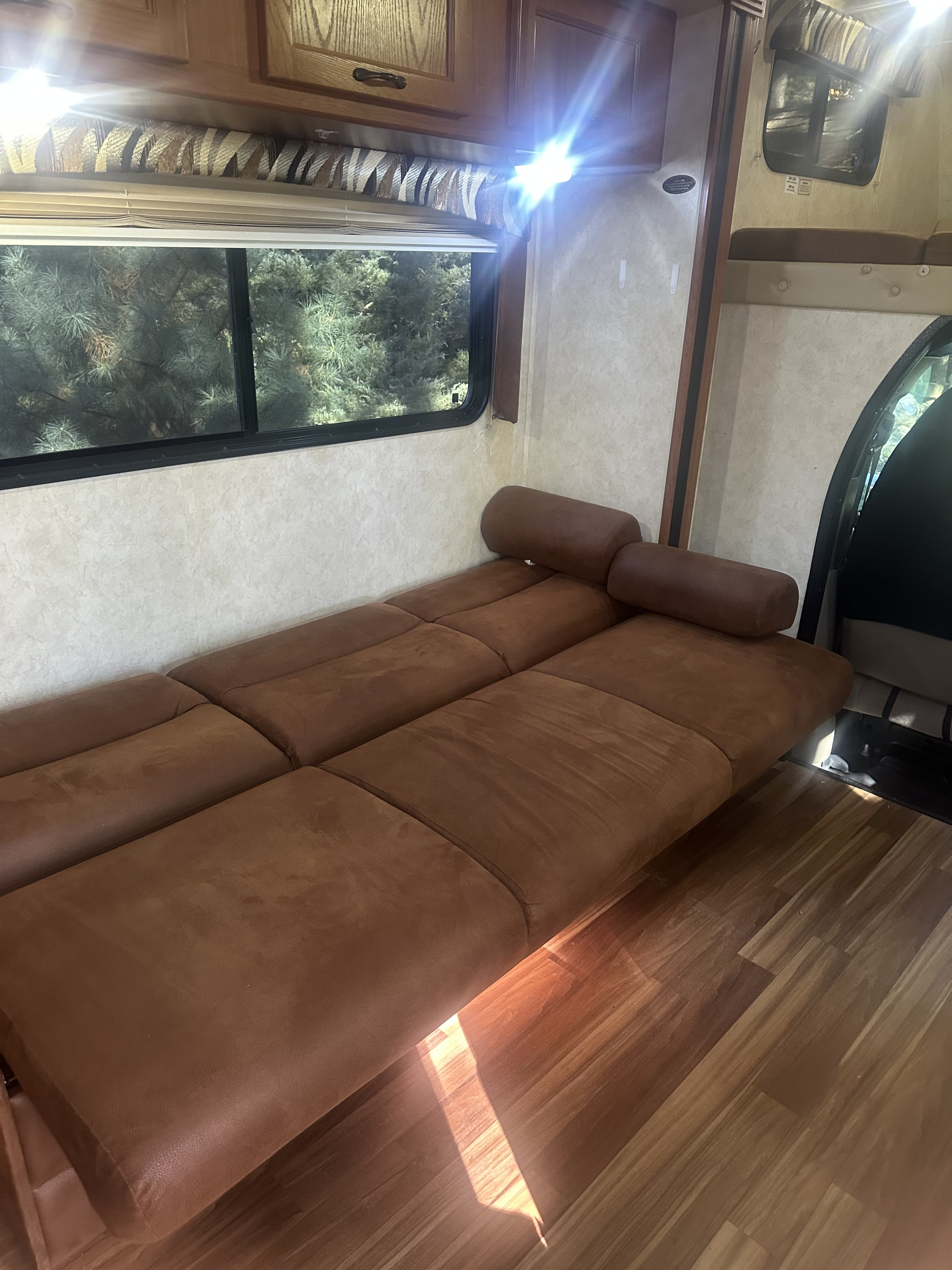 Interior of an RV rental: a brown sectional sofa, window with trees outside, wood-look floor, and bright overhead lights.. Coachmen Coachmen Freelander 2013