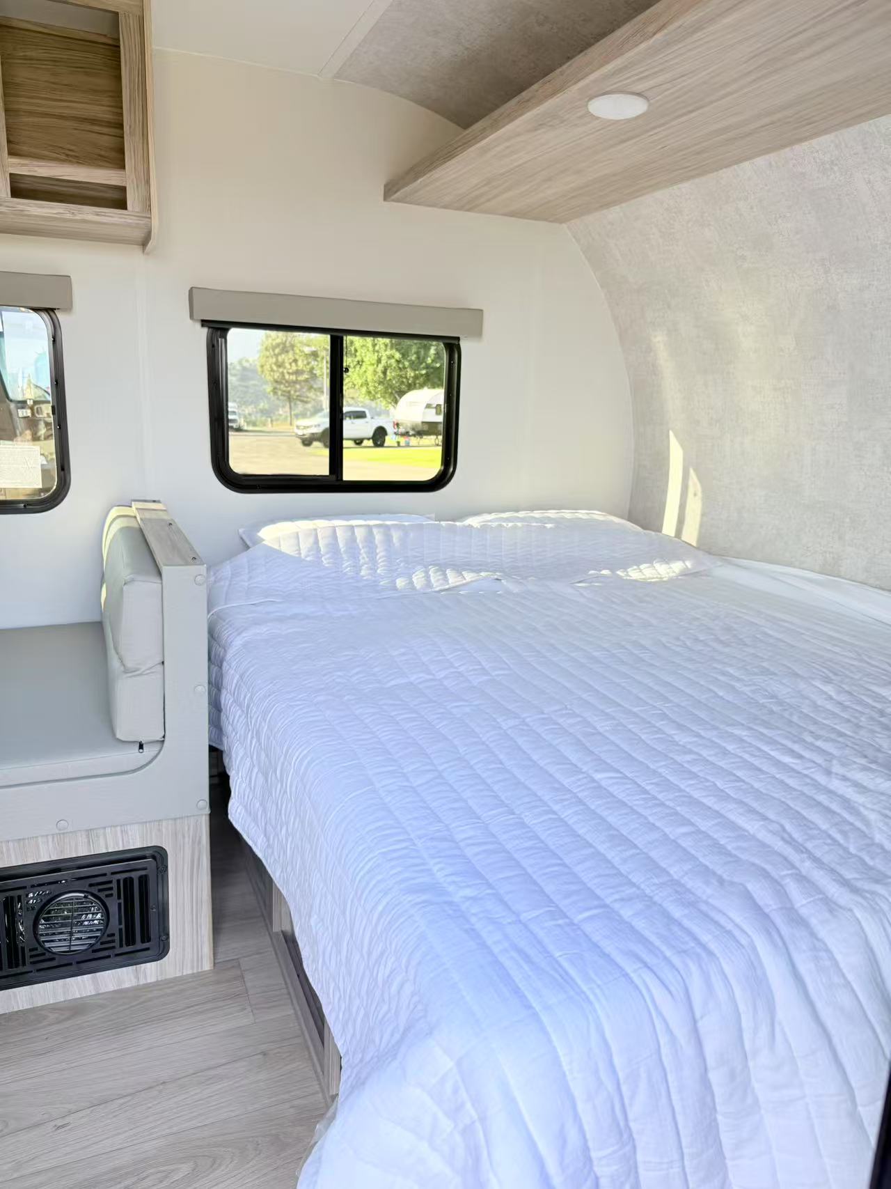 Bright rv rental interior featuring a white-quilted bed, light wood accents, and windows looking out to a campsite.. Winnebago Access 15BH 2025