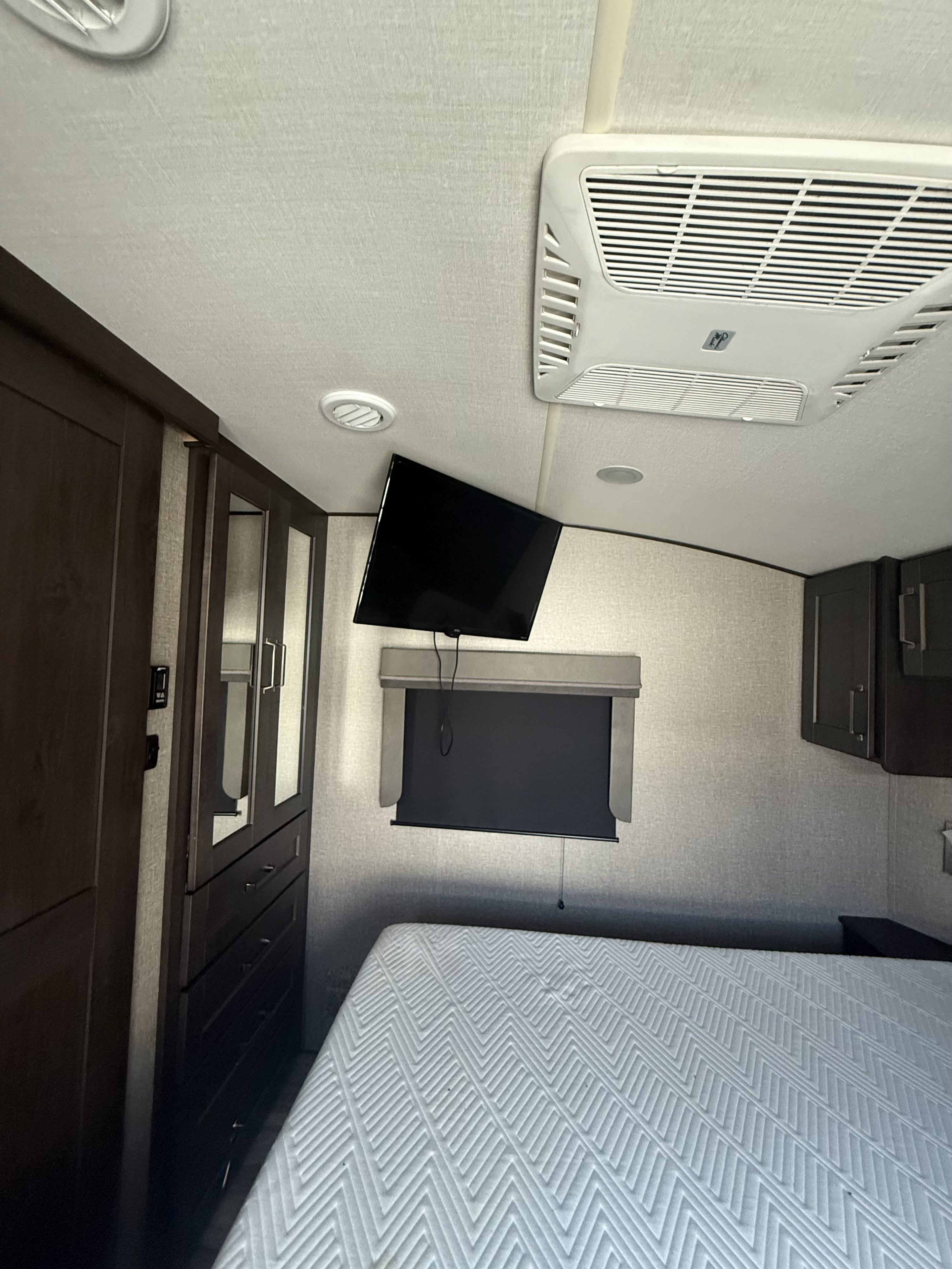 RV rental bedroom interior, featuring a bed with a white patterned mattress, wall-mounted TV, AC unit, and built-in dark wood cabinets.. Grand Design Reflection 2021
