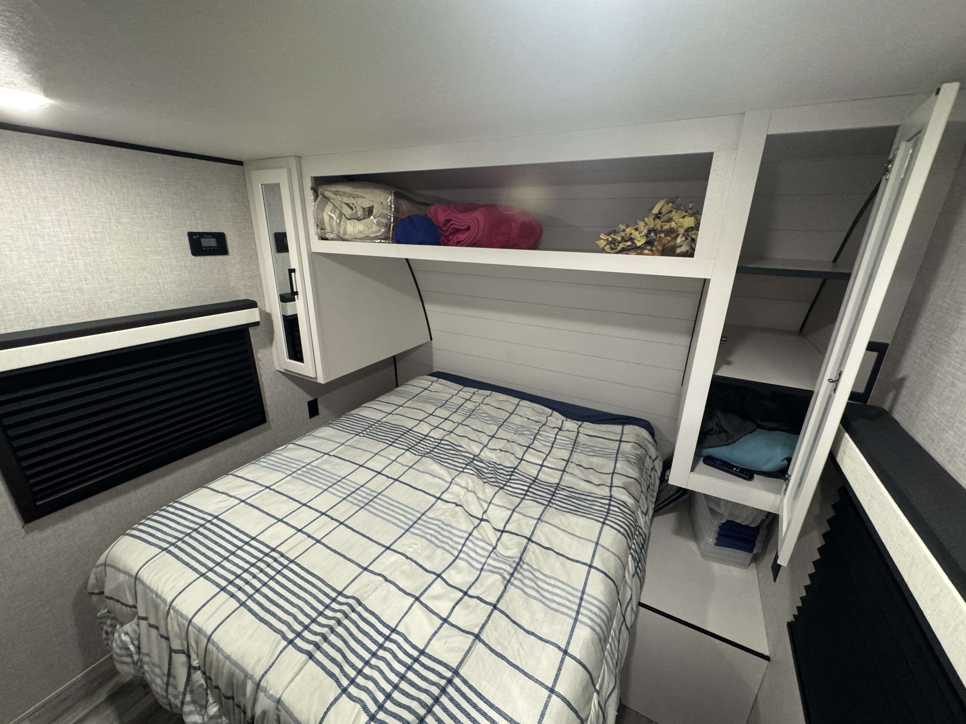 Compact RV rental bedroom with a plaid bed, overhead shelving, open storage, and a wall control panel.. Jayco Jay Flight 2024
