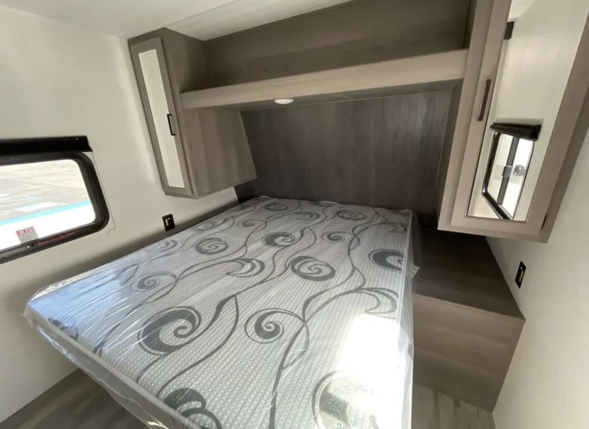 Cozy RV rental bedroom with a new, plastic-covered bed, overhead cabinets, and a bedside window for comfortable travels.. Keystone RV Hideout Sport 2024