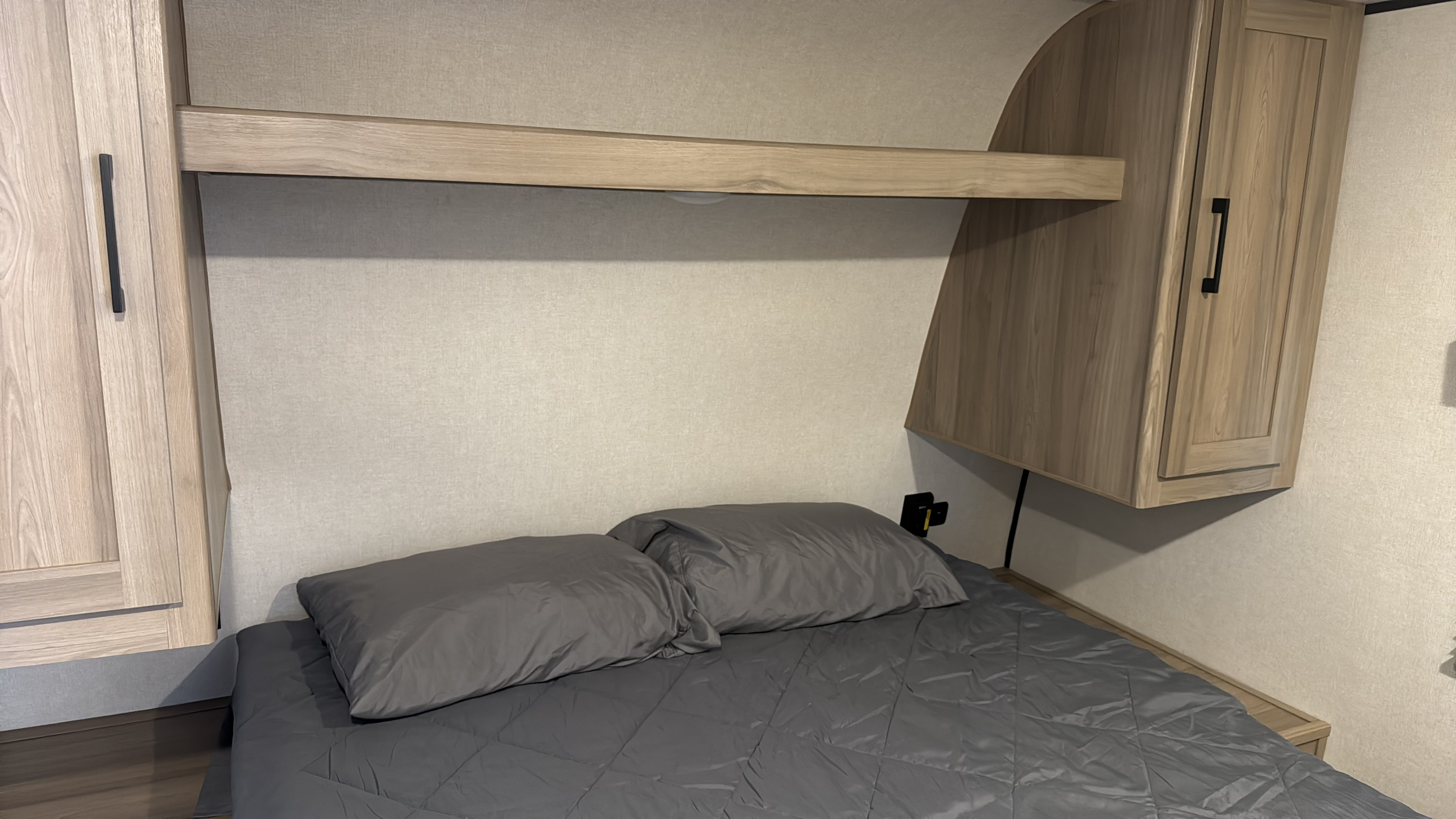 Comfortable RV rental bedroom featuring a grey bed, overhead shelf, and light wood-tone storage cabinets on each side.. Keystone Coleman 22BX 2025