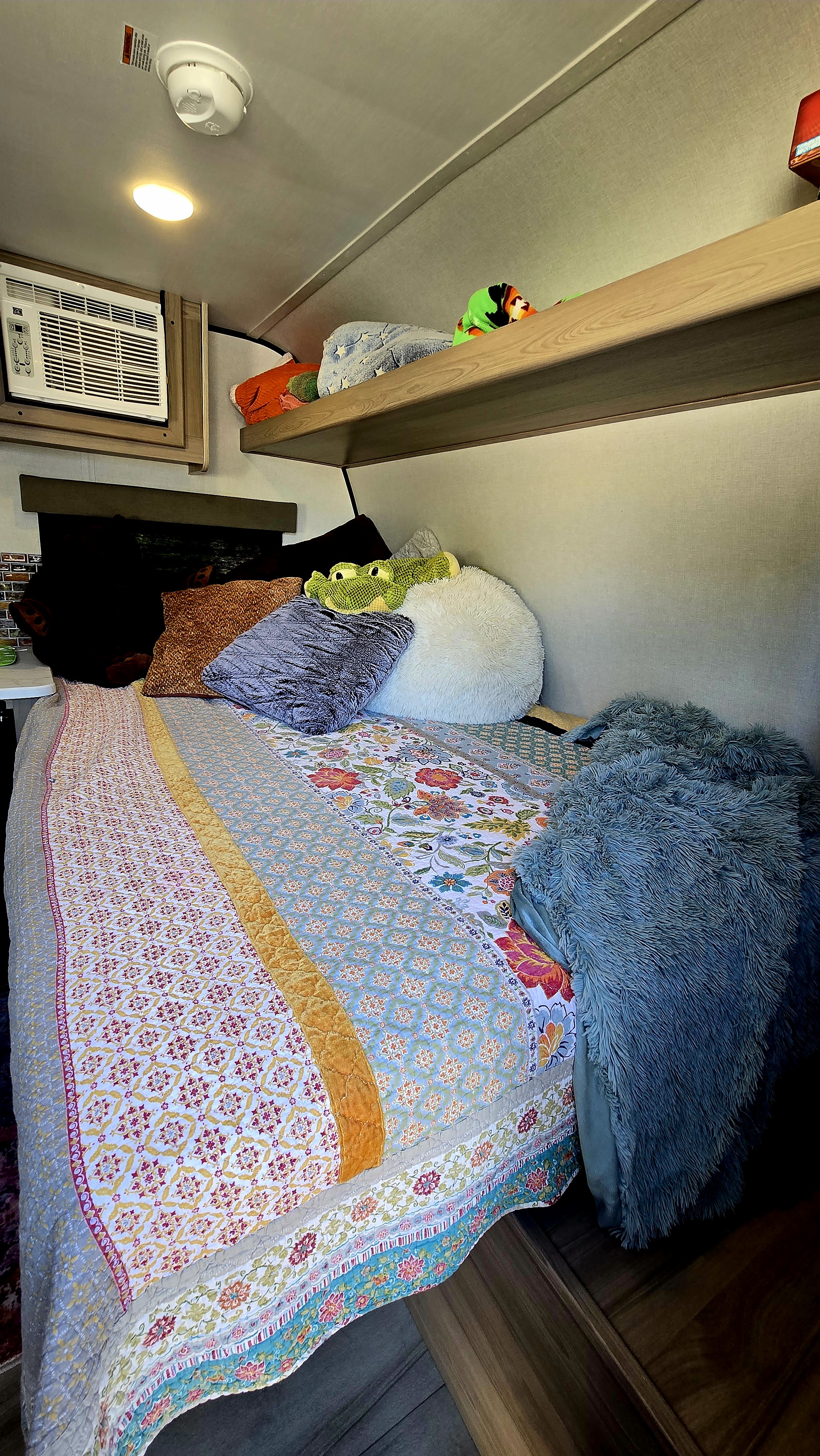 Cozy RV rental bedroom features a vibrant quilt, plush pillows, AC, and an overhead shelf for a comfortable stay.. Keystone 13r 2026