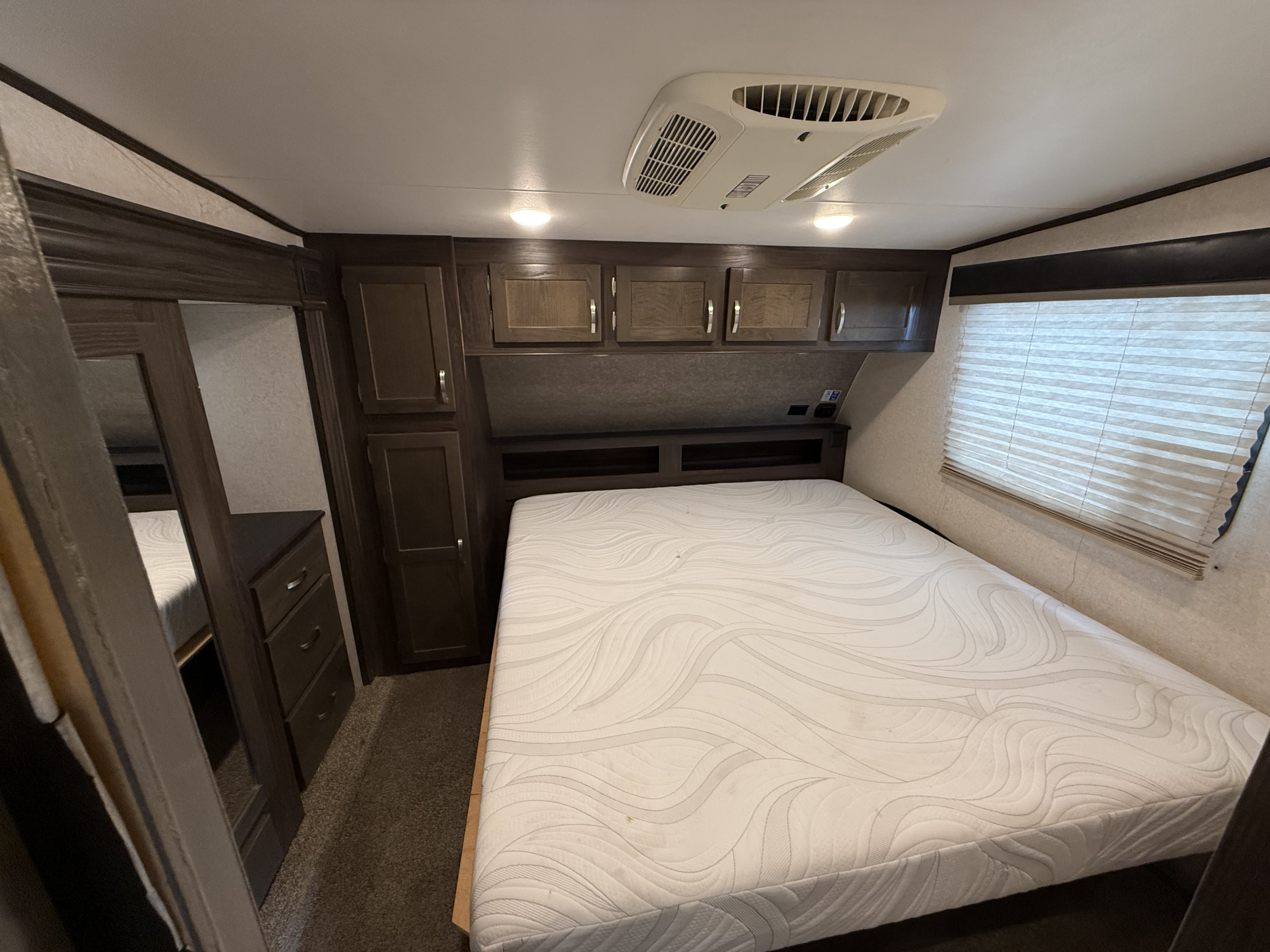 Cozy RV rental bedroom showing a bed, dark wood cabinets, AC unit, and a window with blinds.. Jayco Octane 2019