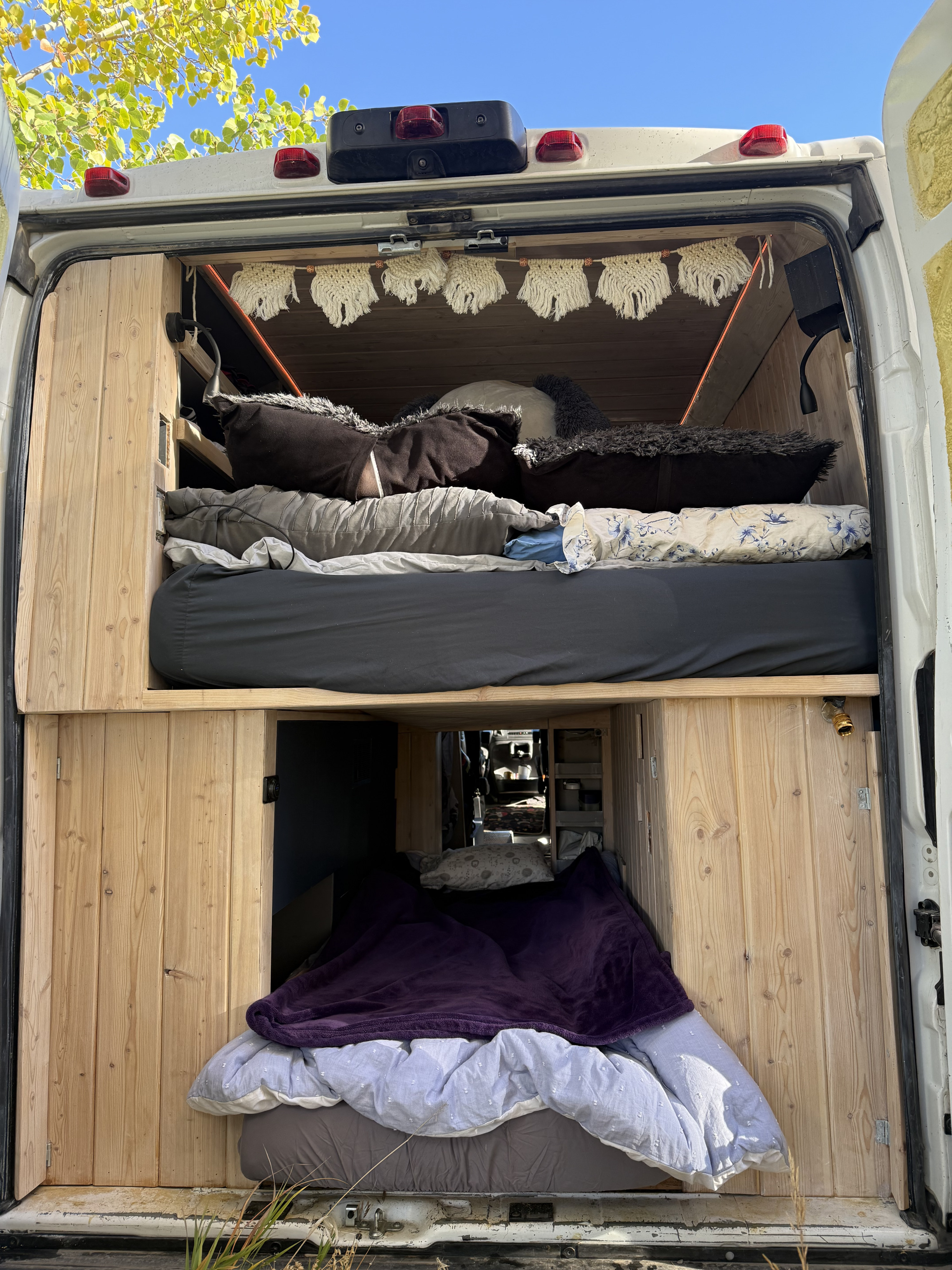 Inside an RV rental van, showing stacked beds, pillows, blankets, and warm wood paneling, viewed from the open rear doors.. Dodge 159 3500 2022