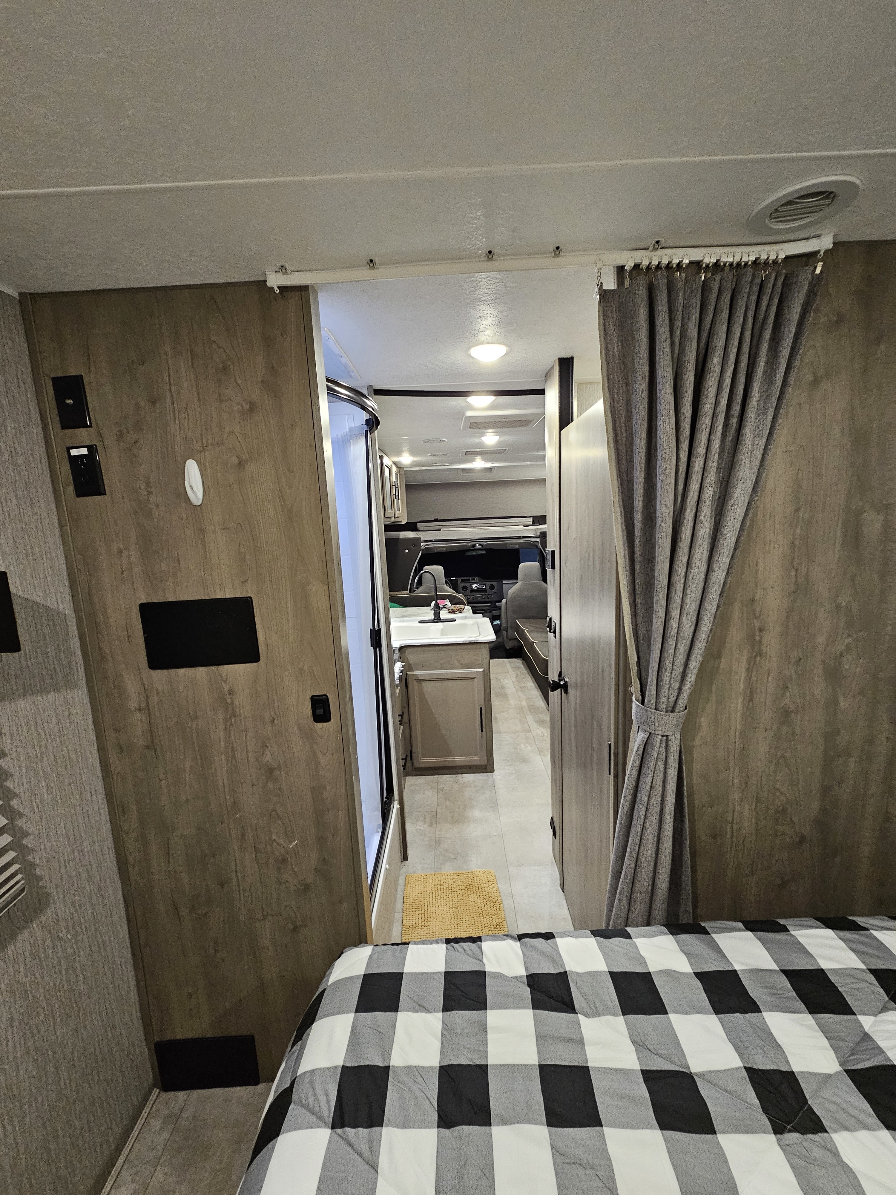 Inside an RV rental: checkered bed, hallway leading past a shower and vanity to the driver's cab area.. Coachmen Freelander 27QB 2022