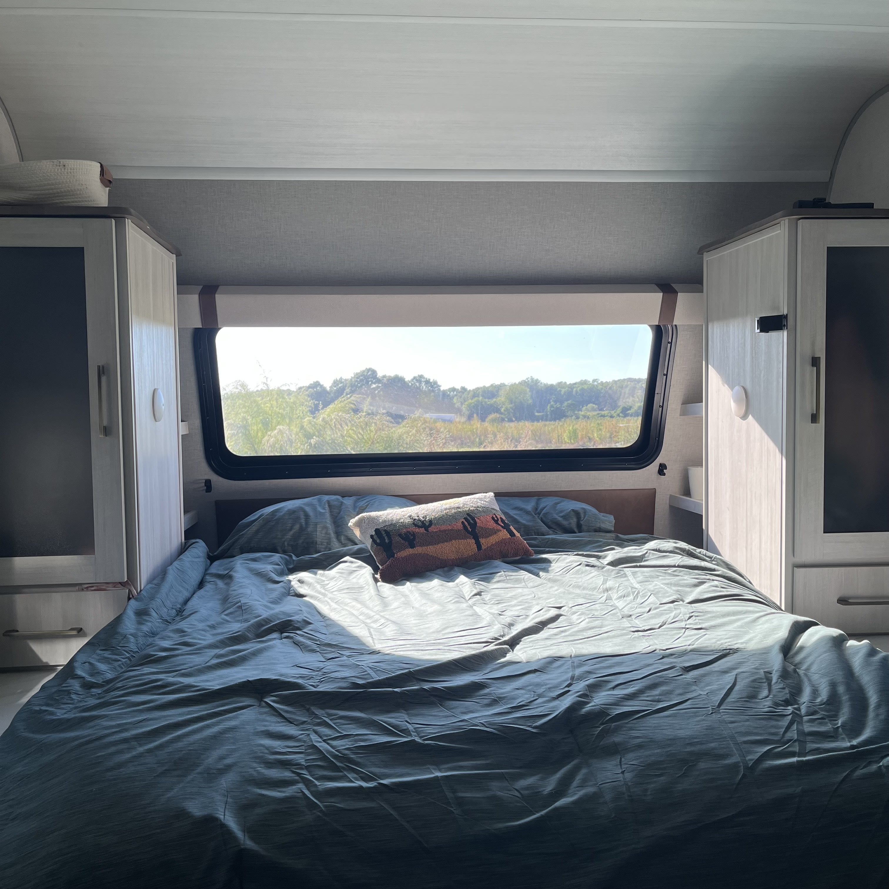Sunlit RV rental bedroom with a comfy bed, desert-themed pillow, window view of trees, and light cabinets.. Forest River 5NHTRDV27TD211453 2026